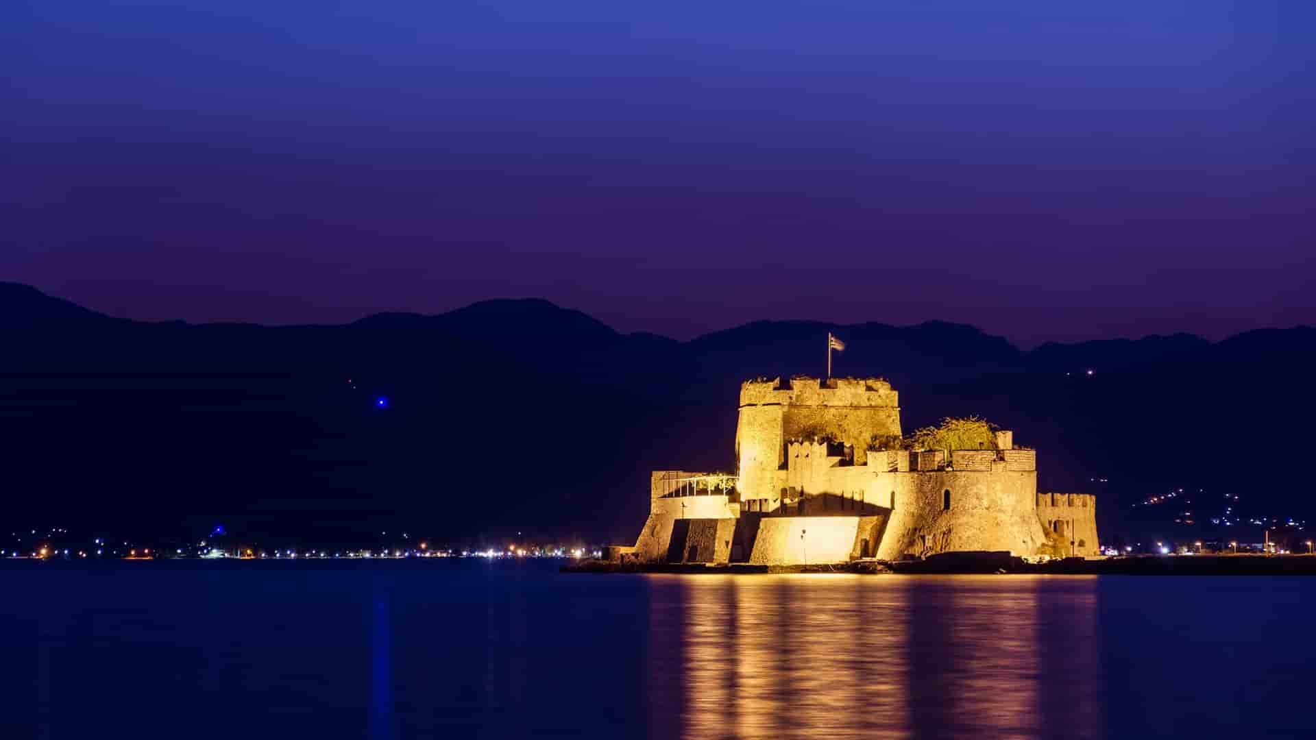 A nighttime long-exposure shot of the Venetian fortress of Bourtzi Castle in Nafplion, Greece, lit up against the dark water and mountains, with city lights in the distance.