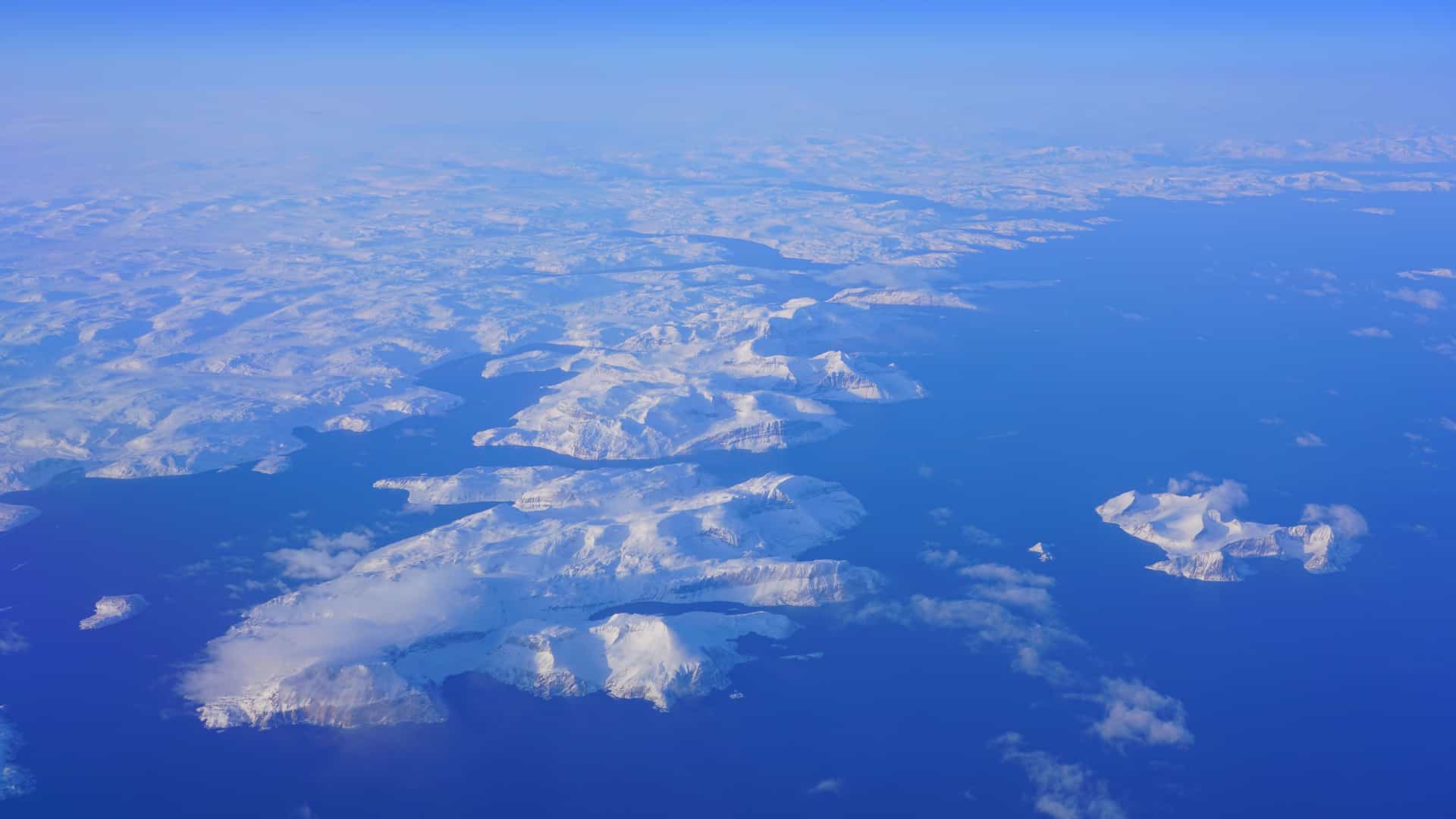 An aerial view of the snow-covered mountains and islands along the coast of Nain, Canada, with vast blue water and a distant shoreline under a clear sky.