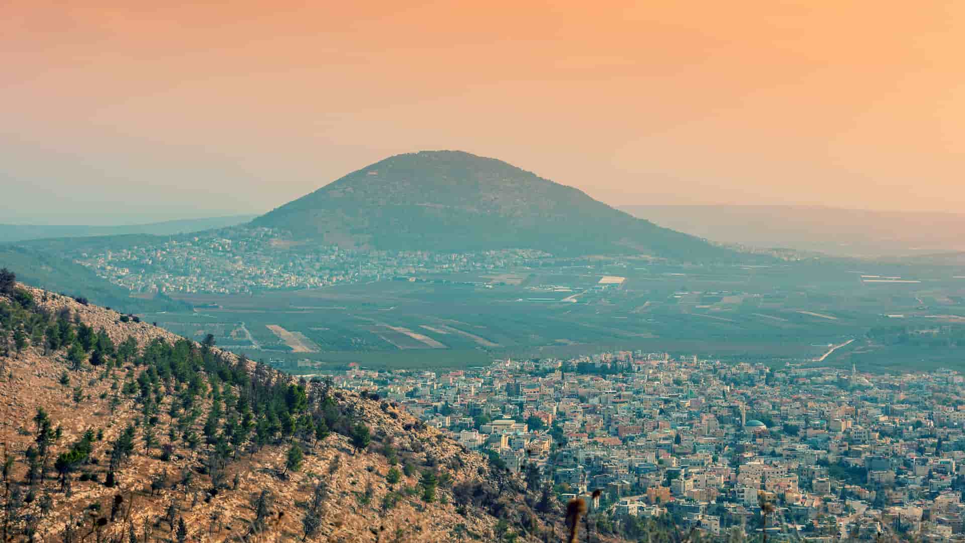 "A high-angle view of the city of Nazareth, Israel, nestled in a valley with Mount Tabor and the surrounding landscape in the distance at sunset. "
