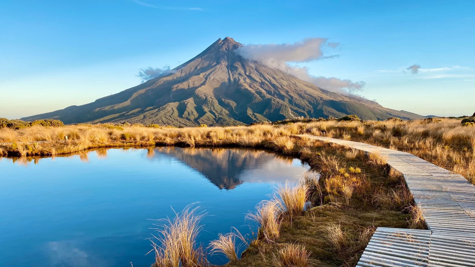A beautiful reflection of the volcanic Mount Taranaki in a calm lake near New Plymouth, New Zealand, with a wooden boardwalk in the foreground.