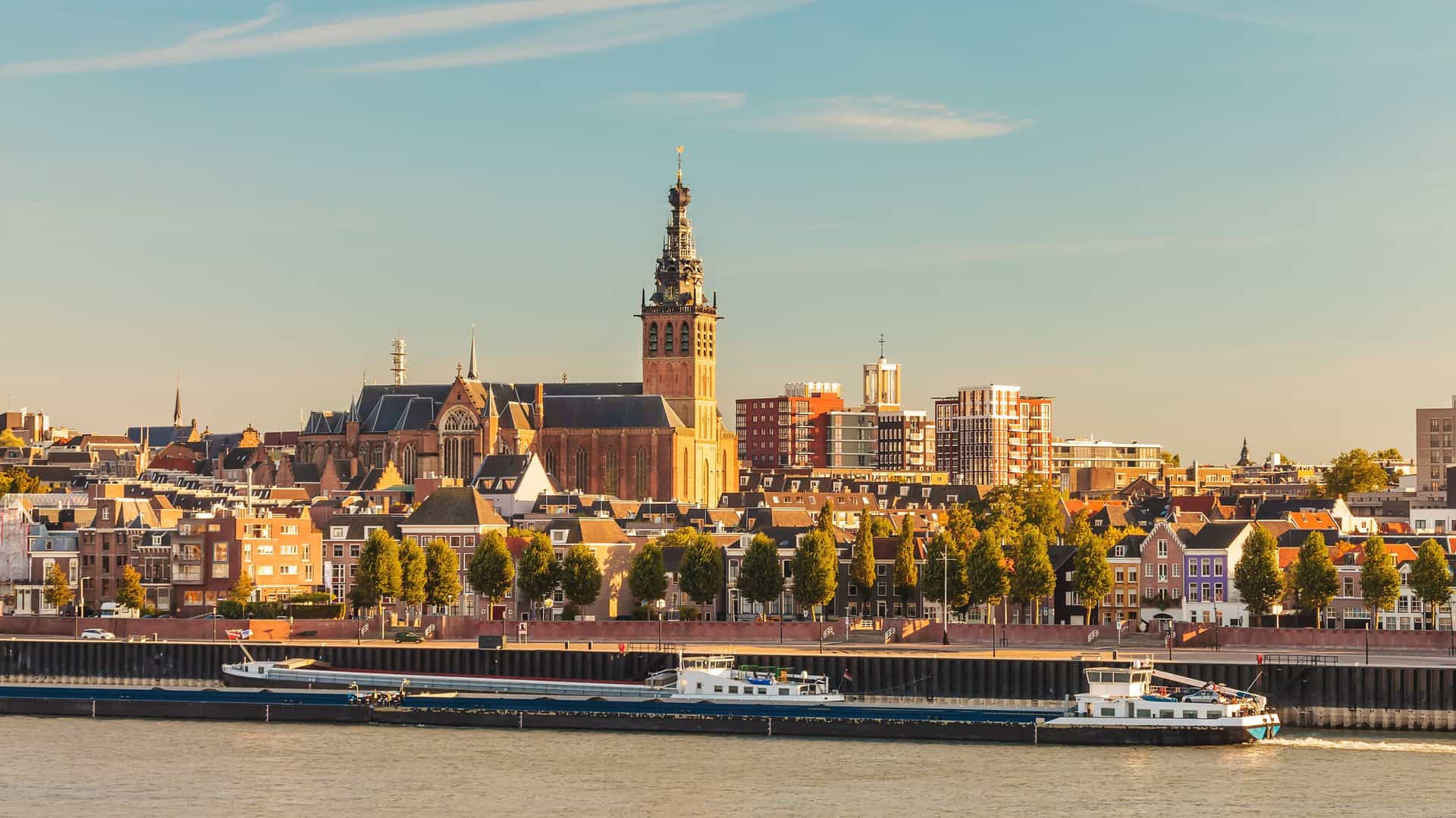 A river cruise barge navigates the Waal River with a panoramic view of the Nijmegen, The Netherlands skyline and the historic Stevenskerk church dominating the cityscape.