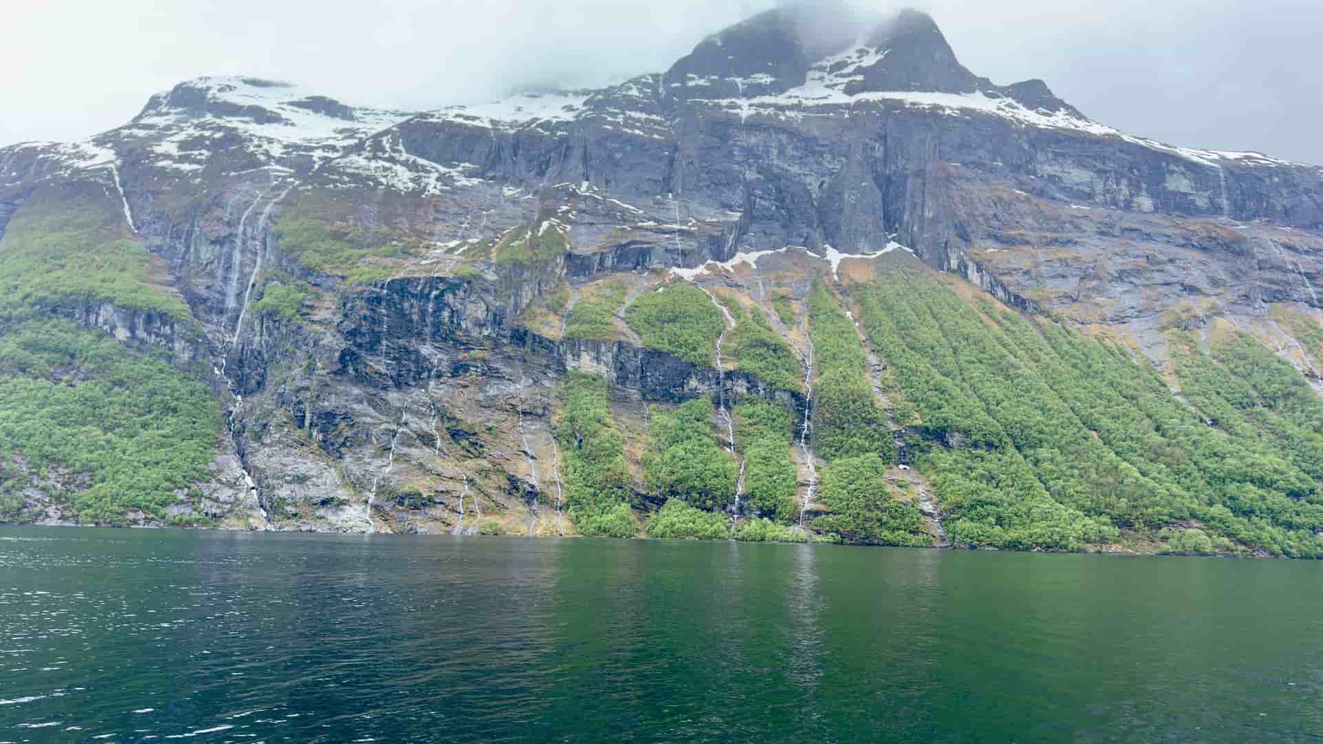 A large cruise ship navigates the calm waters of the Nordfjord in Norway, with a breathtaking view of the steep, rocky cliffs and a large waterfall cascading down into the fjord below.