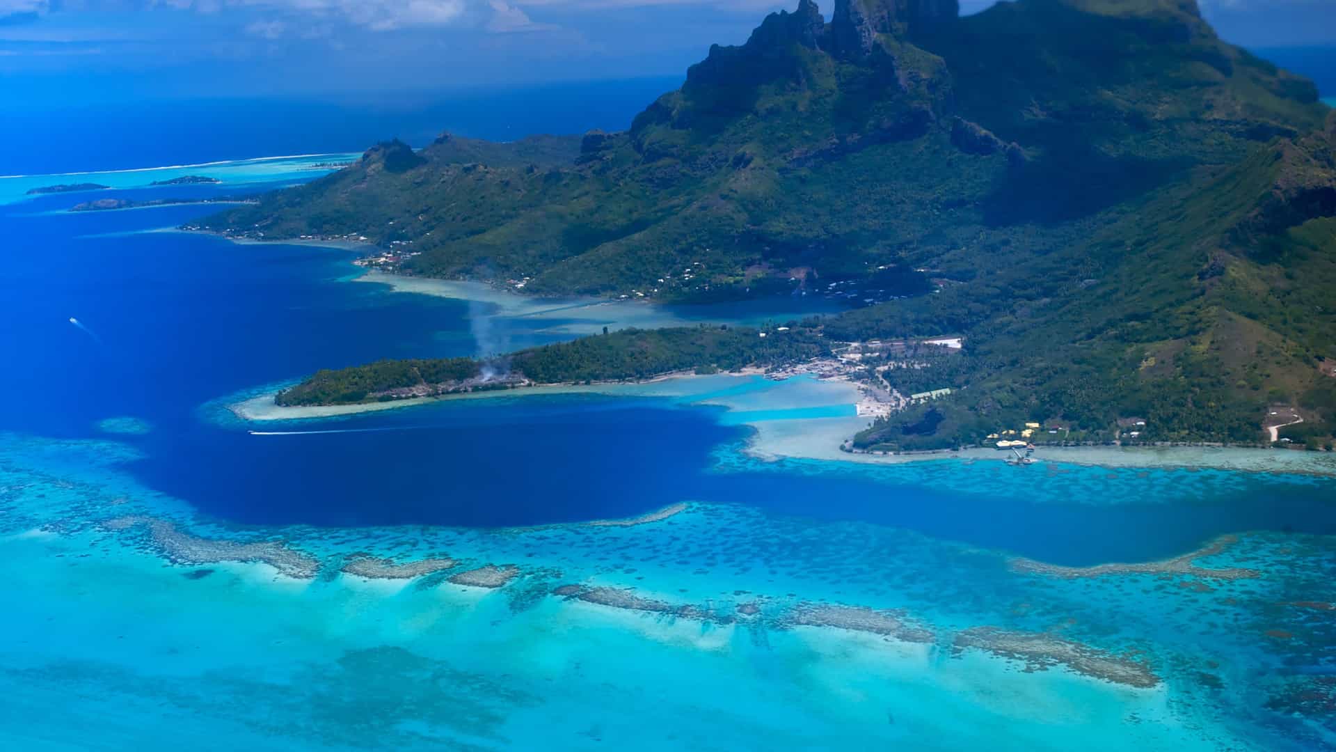 An aerial view of Nuku Hiva in the Marquesas Islands, French Polynesia, showing a volcanic island surrounded by a vibrant turquoise lagoon and coral reefs.