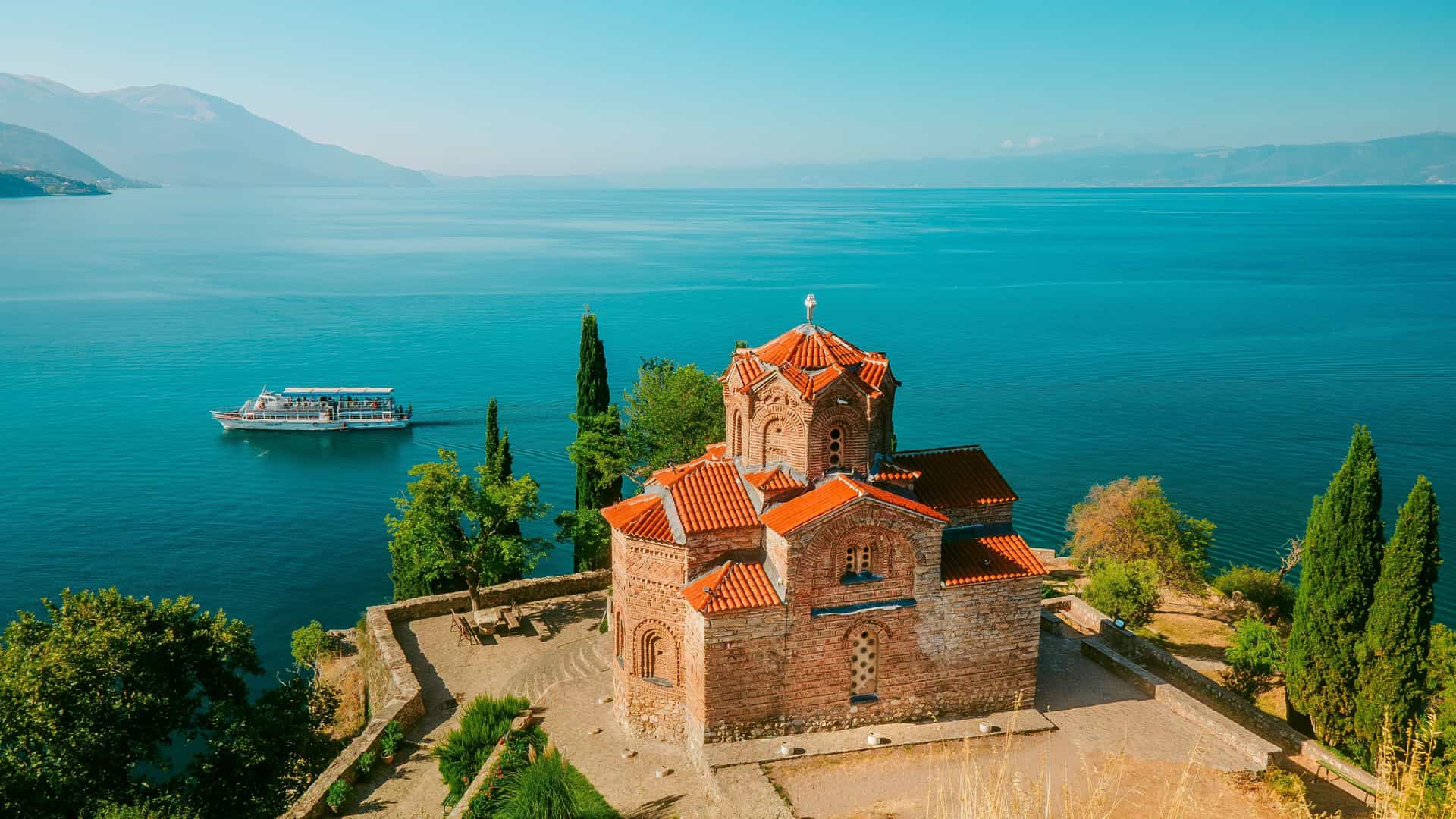 "A panoramic view of the historic Church of St. John the Theologian at Kaneo, overlooking the serene blue waters of Lake Ohrid in Ohrid, North Macedonia, with a tour boat in the background. "