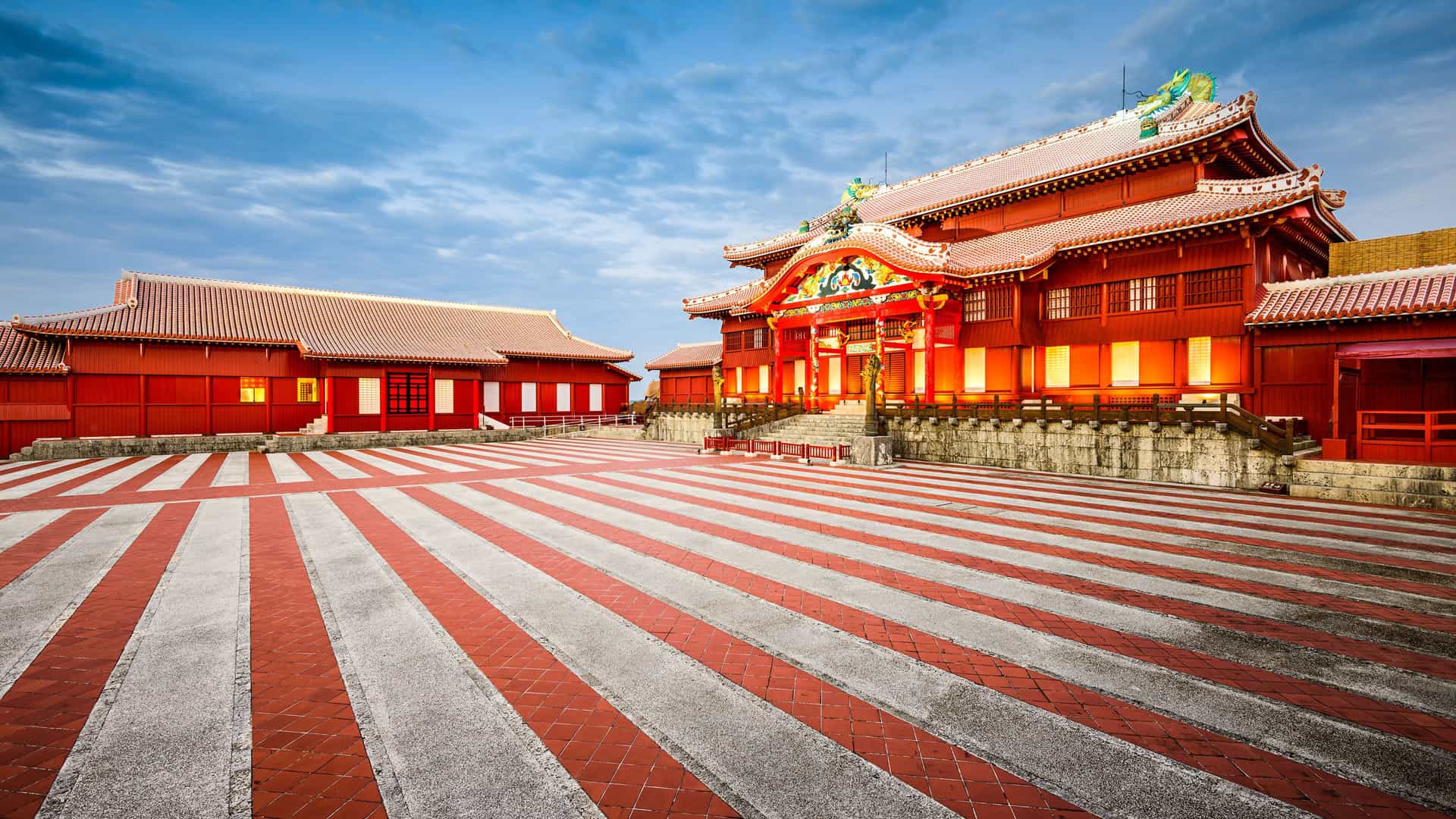 A stunning daytime view of the vibrant red buildings of the Shuri Castle grounds in Naha, Okinawa, Japan, with its wide courtyard featuring a distinctive red and white striped pattern.