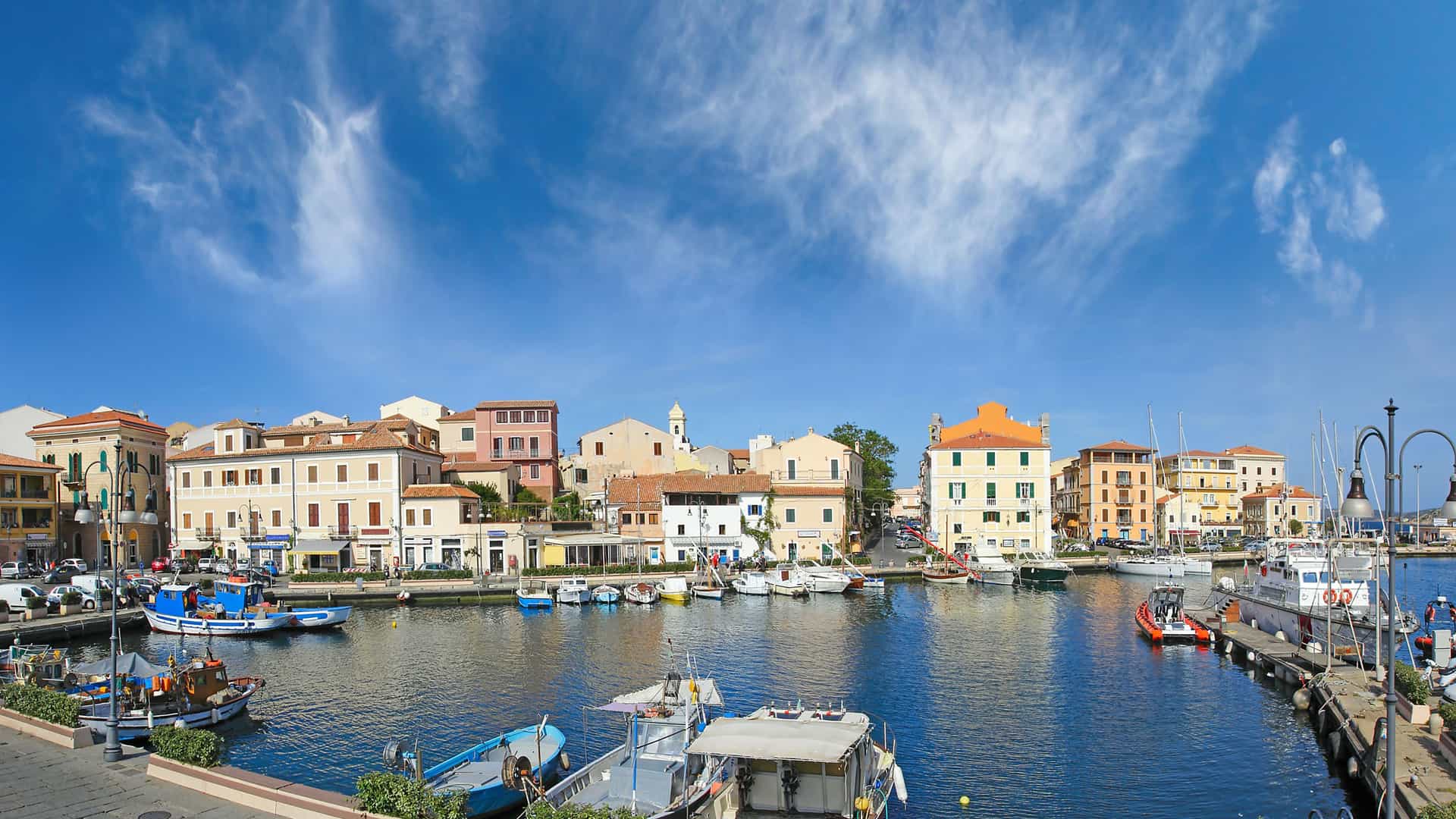 A vibrant panoramic view of the harbor in Olbia, Italy, with a wide variety of fishing boats and yachts docked in front of colorful waterfront buildings and a bright blue sky.