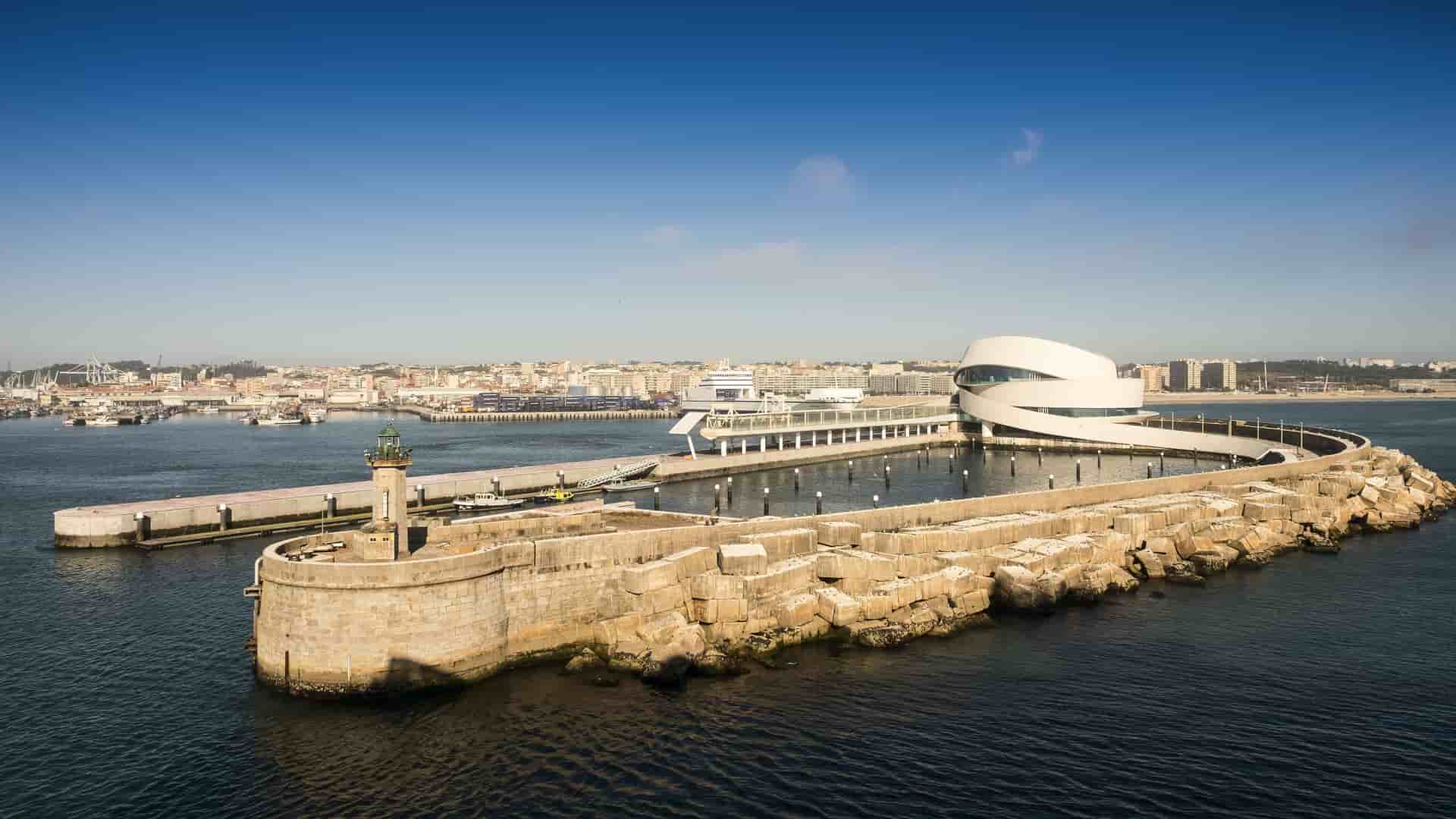 A high-angle view of the modern, white spiral-shaped cruise terminal at the Port of Leixões, Oporto, Portugal, extending into the ocean next to a rocky breakwater.