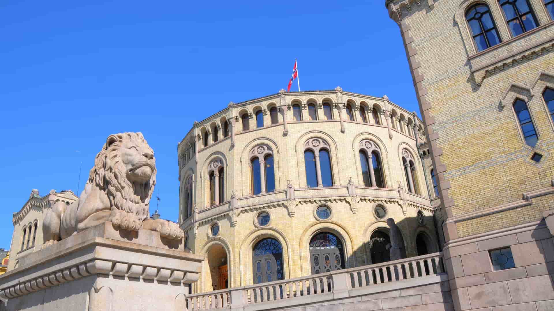A large stone lion sculpture guarding the entrance of the Stortinget, the Parliament of Norway, in Oslo, a historic and prominent landmark against a clear blue sky.