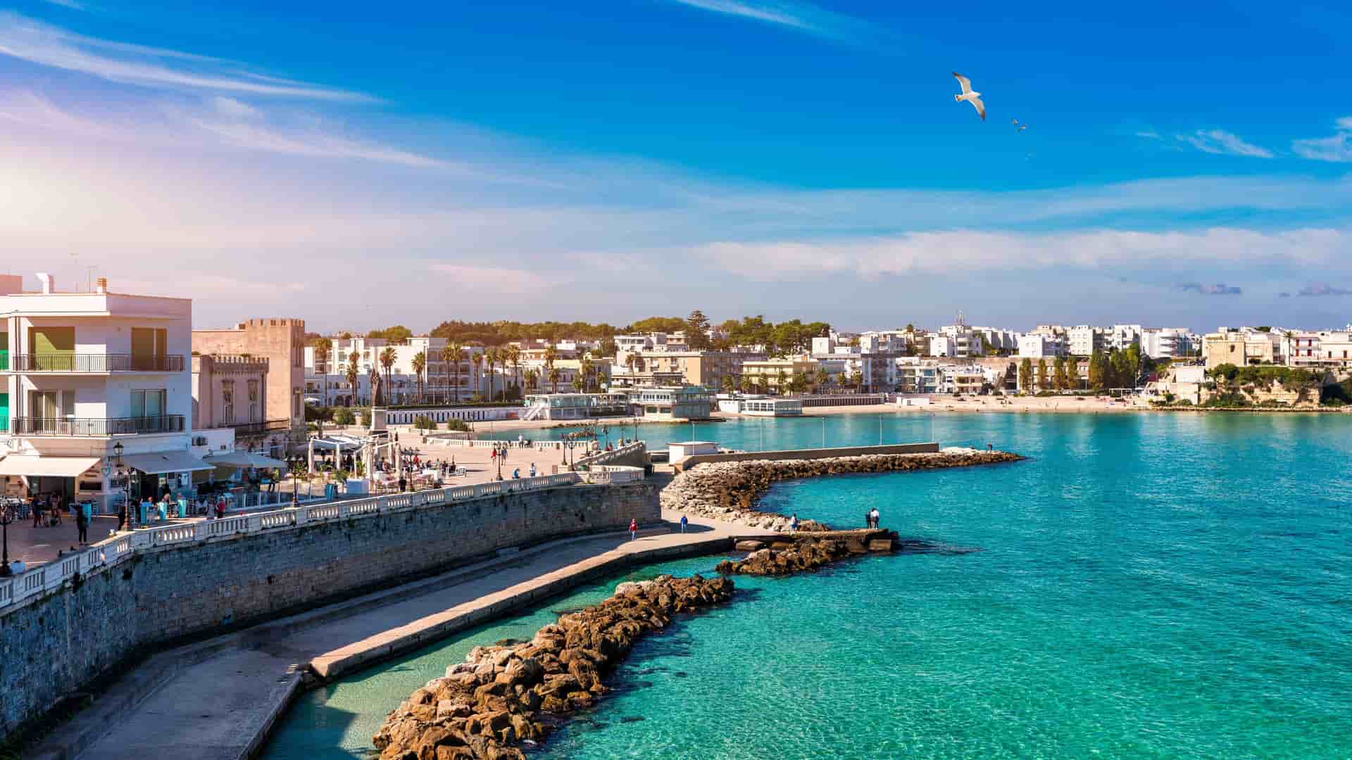 A picturesque view of Otranto, Italy, featuring a promenade along the turquoise-blue sea, with white buildings and a sandy beach under a bright blue sky.