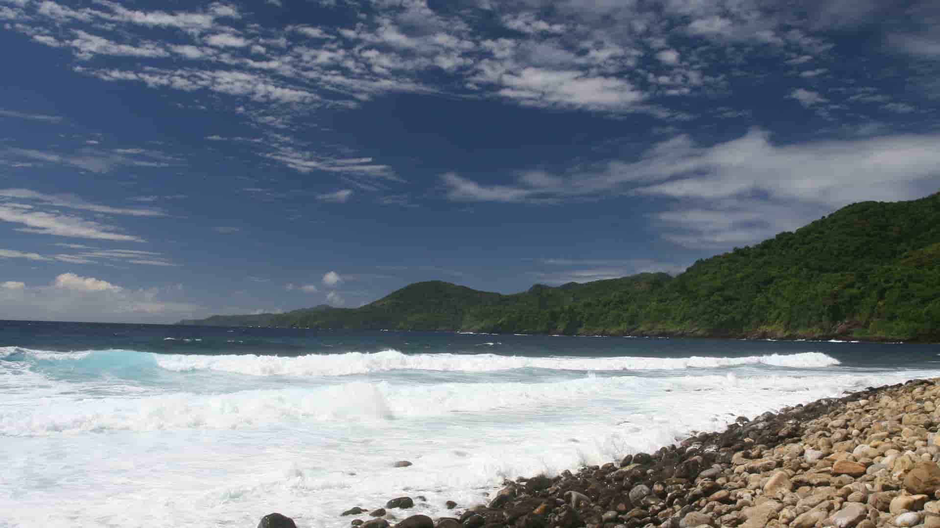 A view of a beautiful beach in Pago Pago, American Samoa, with large waves crashing on a rocky shore, surrounded by lush, green hills.