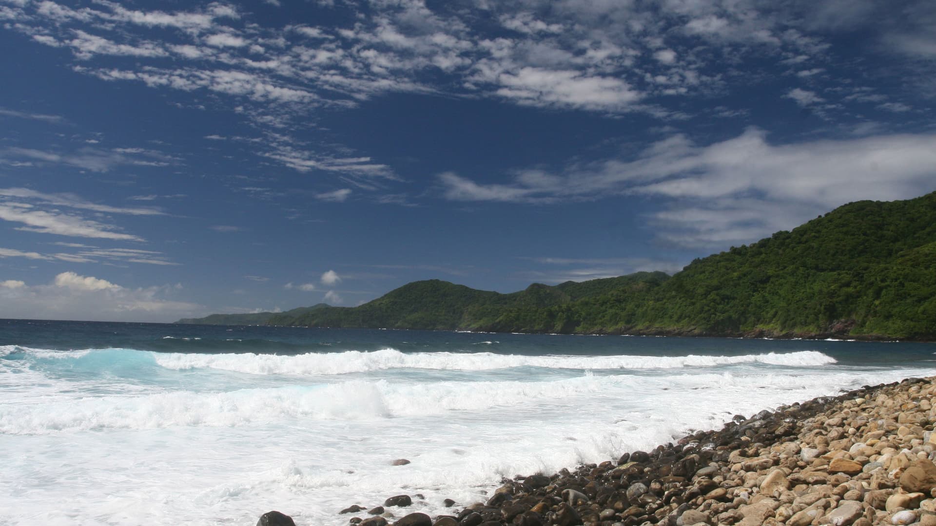 A view of a beautiful beach in Pago Pago, American Samoa, with large waves crashing on a rocky shore, surrounded by lush, green hills.
