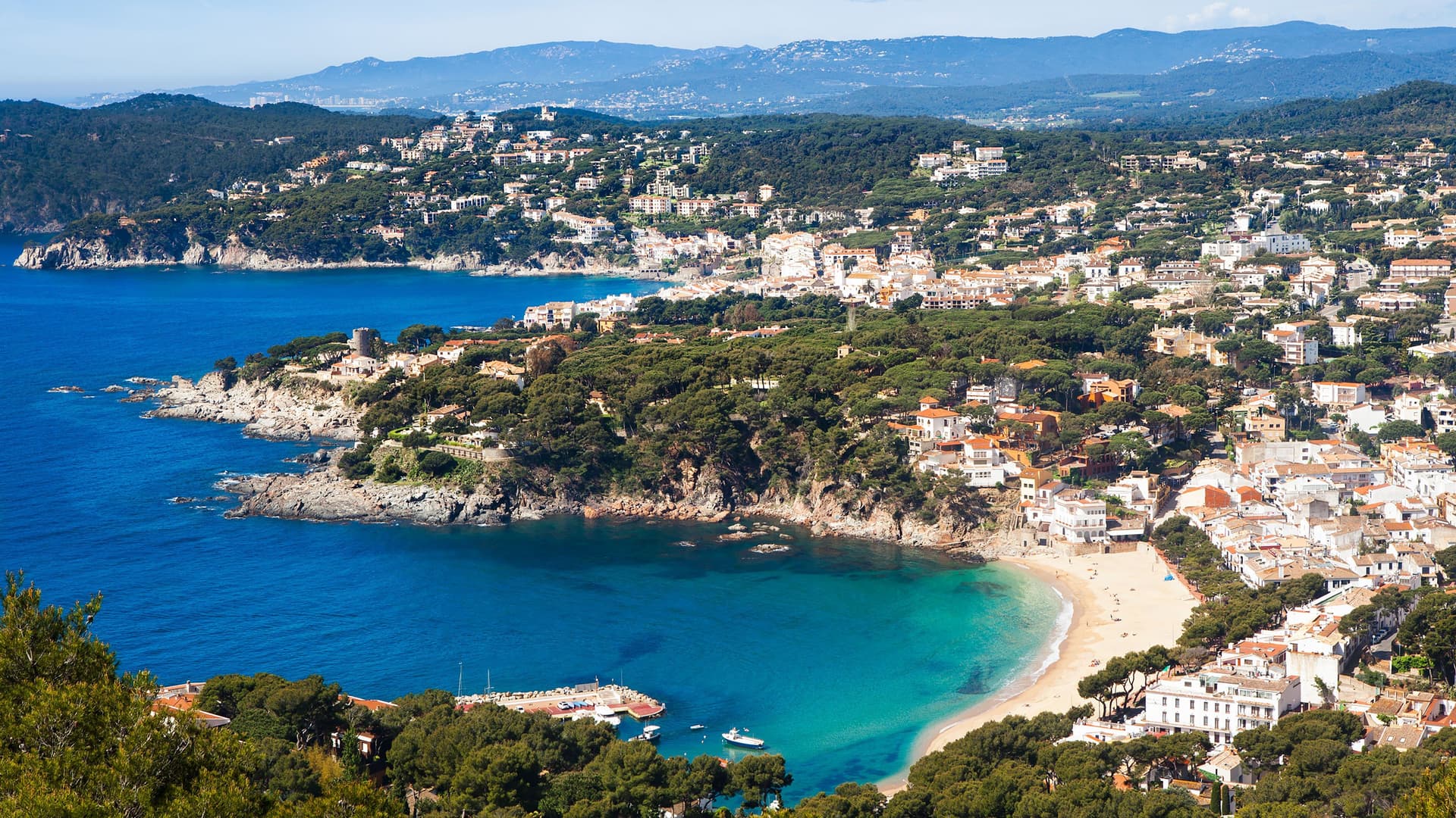 An aerial panoramic shot of the stunning coast of Palamós, Spain, with a beautiful beach, clear turquoise water, and houses built on the hillside.