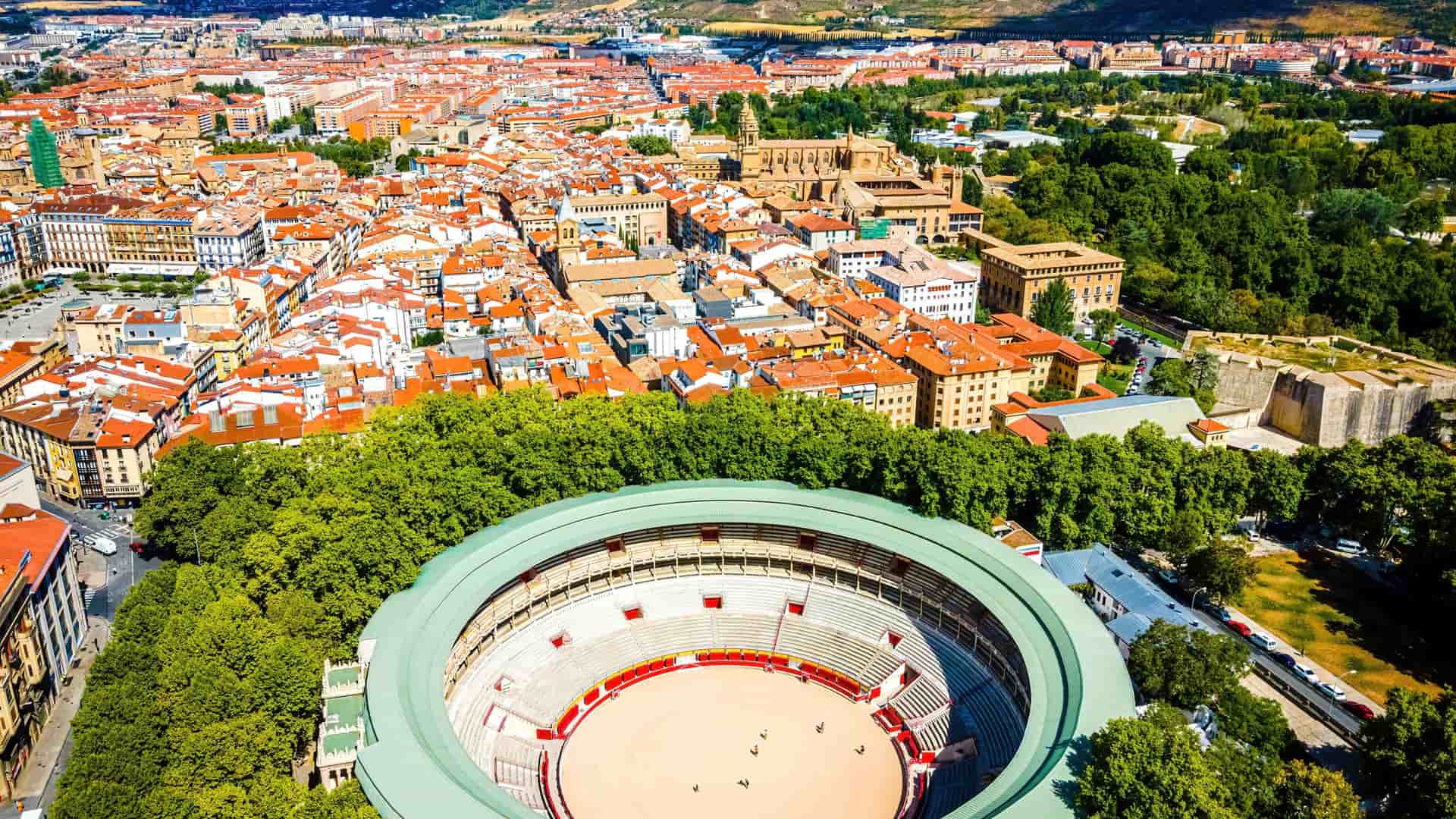 "An aerial panoramic shot of the historic Plaza de Toros, the bullring in Pamplona, Spain, with the city's old town and surrounding green trees and landscape in the background. "