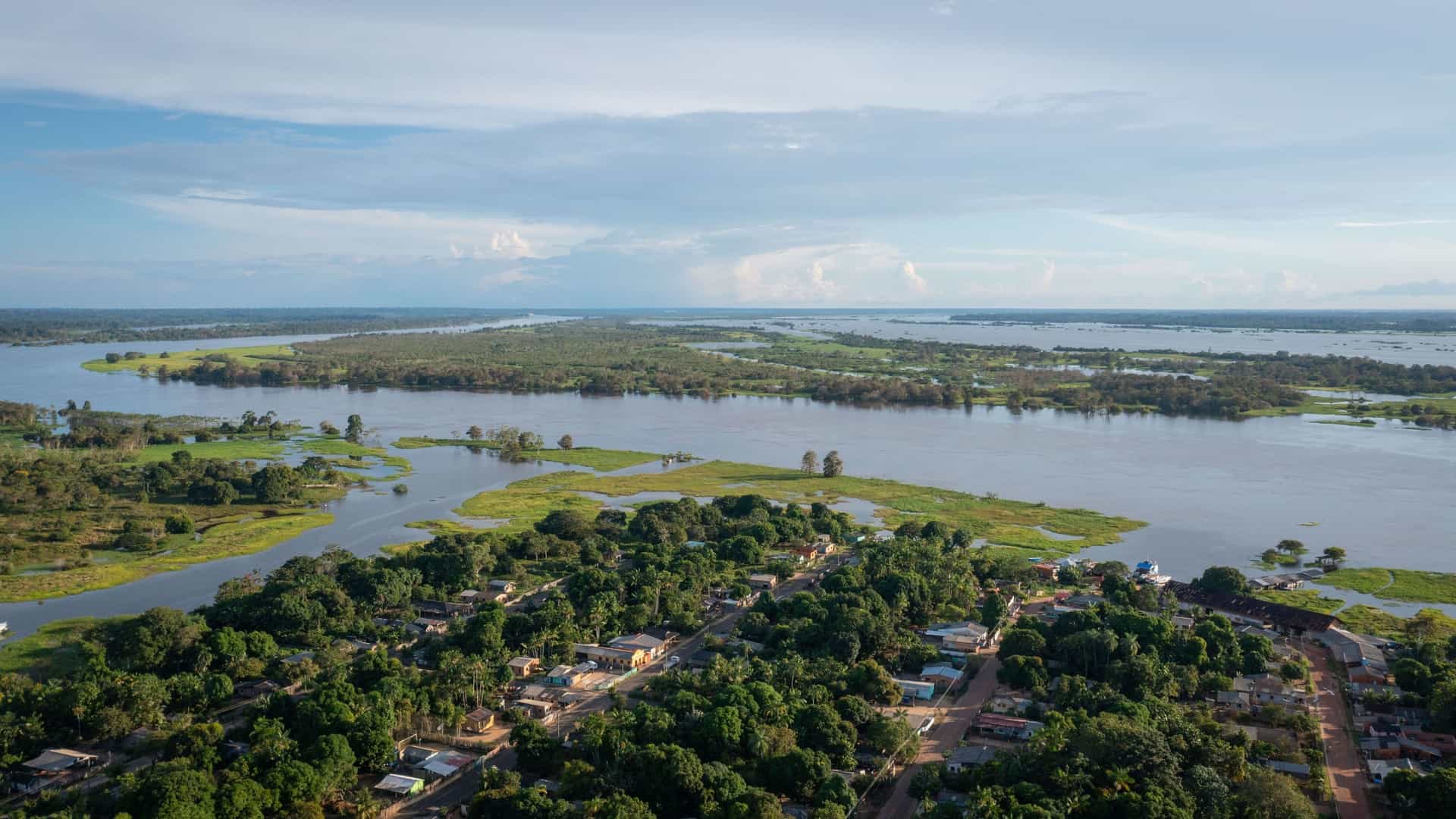 An aerial view of the city of Parintins, Brazil, located on the Amazon River, with a dense forest in the foreground and the wide river in the distance.