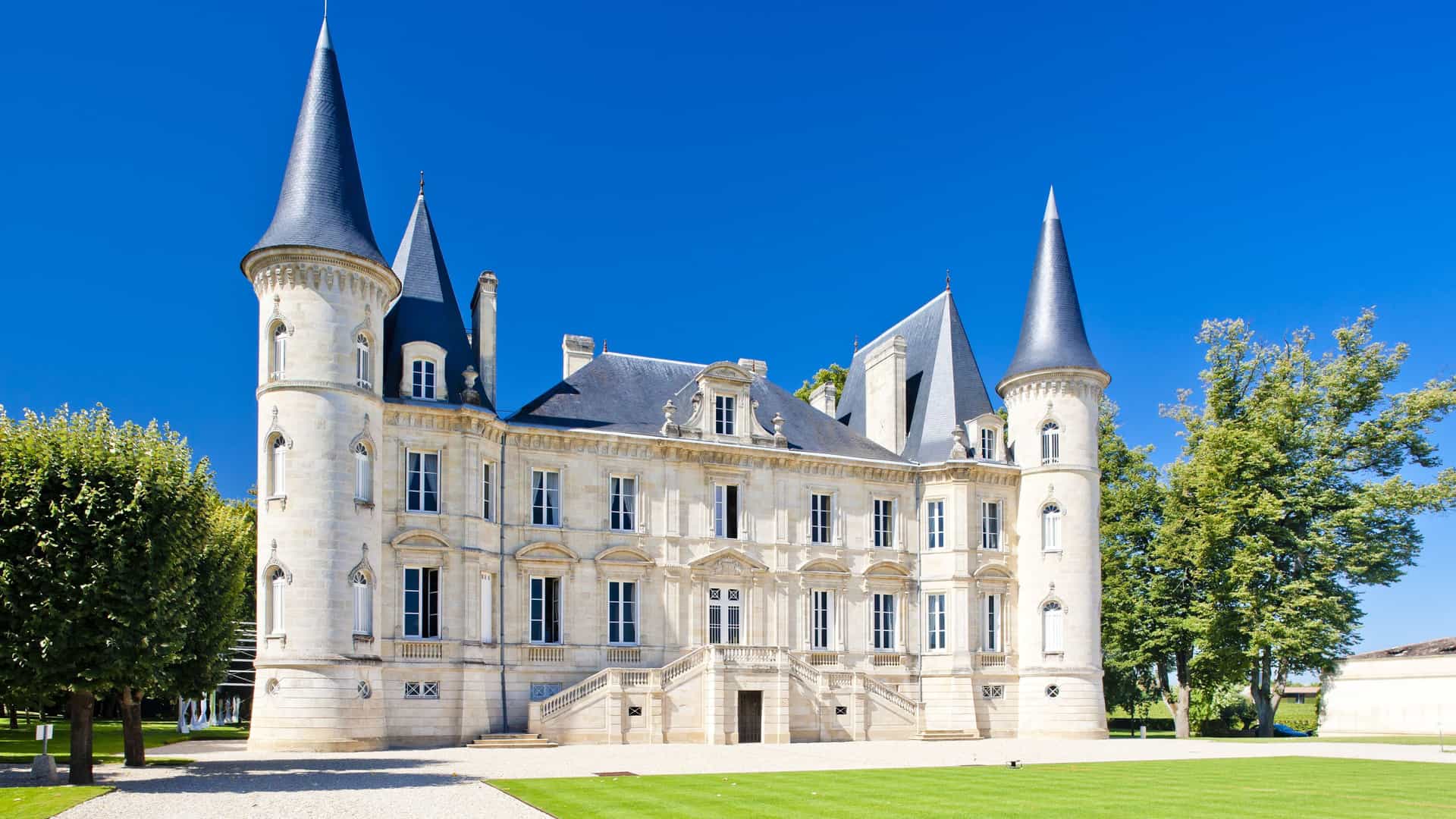 A grand, historic château with multiple turrets and a perfectly manicured lawn, the famous Château Pichon Baron, a beautiful landmark in Pauillac, France.