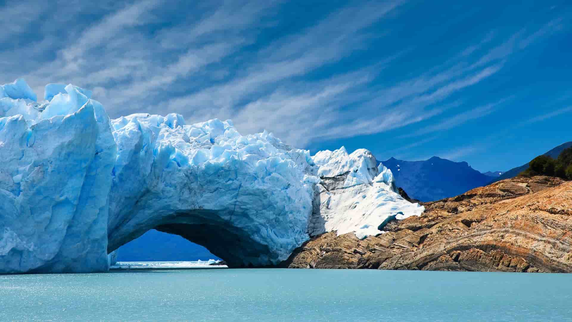 A breathtaking view of the massive Pio XI Glacier in Chile, showing its bright blue ice arching over the water, with a rocky shoreline and mountains in the distance under a blue sky.