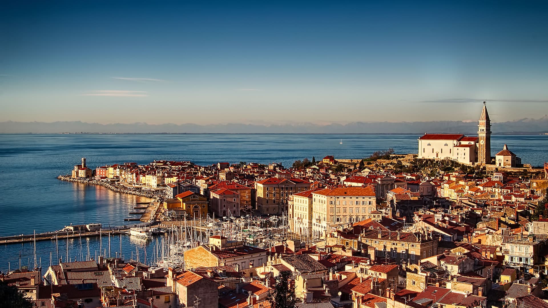 A scenic, high-angle view of the historic coastal town of Piran, Slovenia, with its densely packed terracotta roofs, a marina filled with boats, and a church with a bell tower overlooking the Adriatic Sea at sunset.