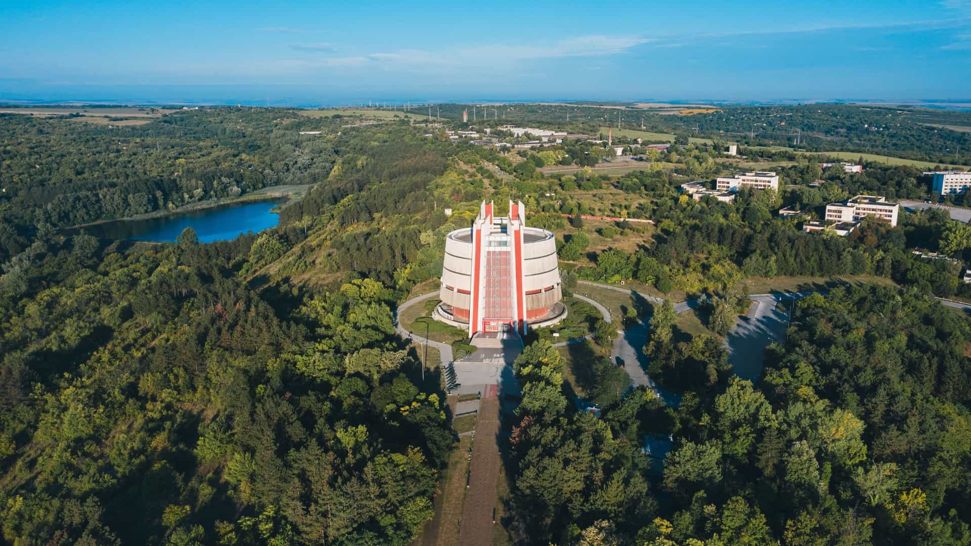 An aerial view of the iconic Pleven Panorama in Bulgaria, a modern, cylindrical building surrounded by dense green forests and a lake, commemorating the Siege of Pleven.