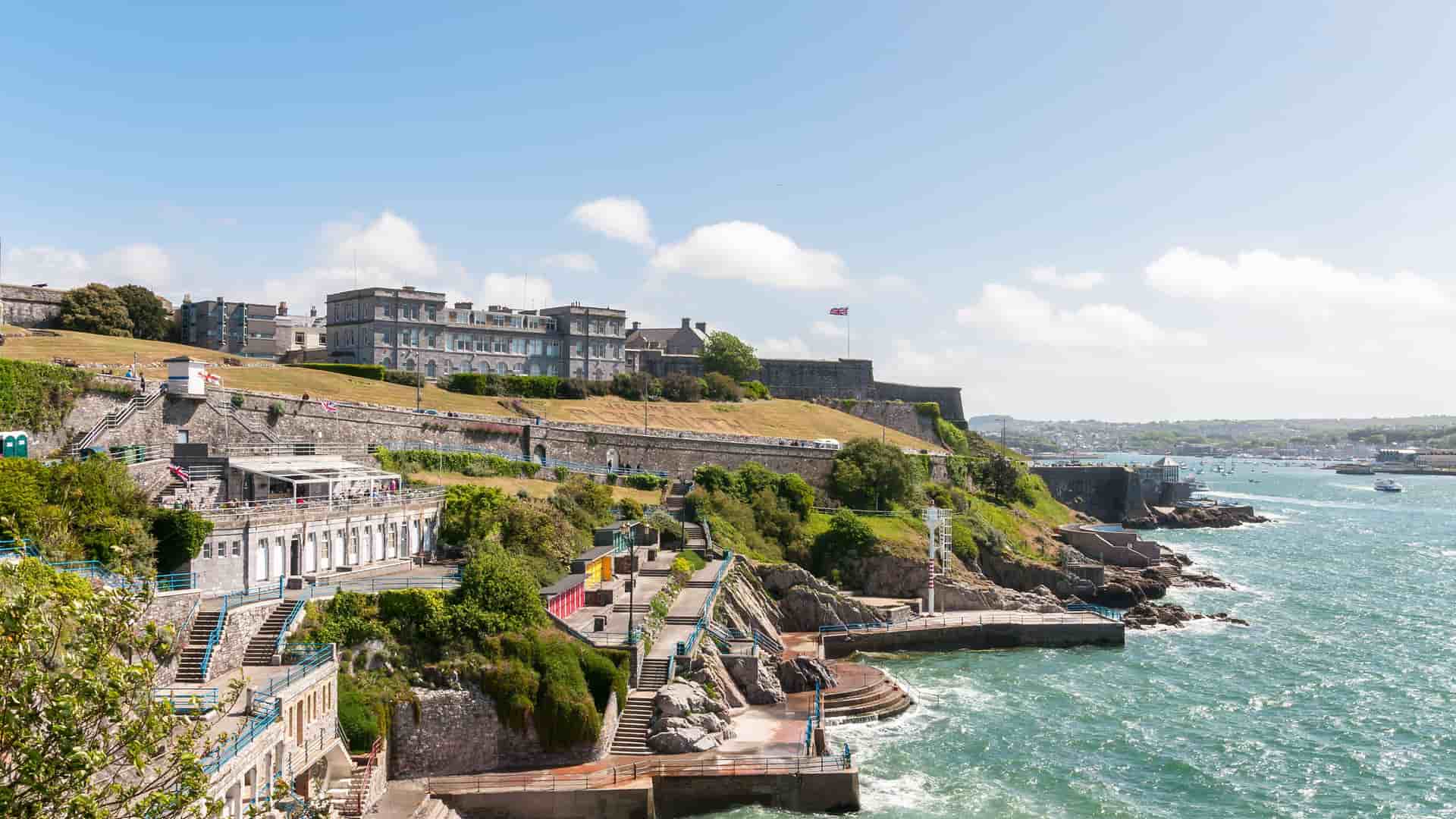 A wide view of Plymouth Hoe, showcasing the historic Citadel, a large grassy area, and the scenic waterfront with views of the bustling harbor in Plymouth, UK.