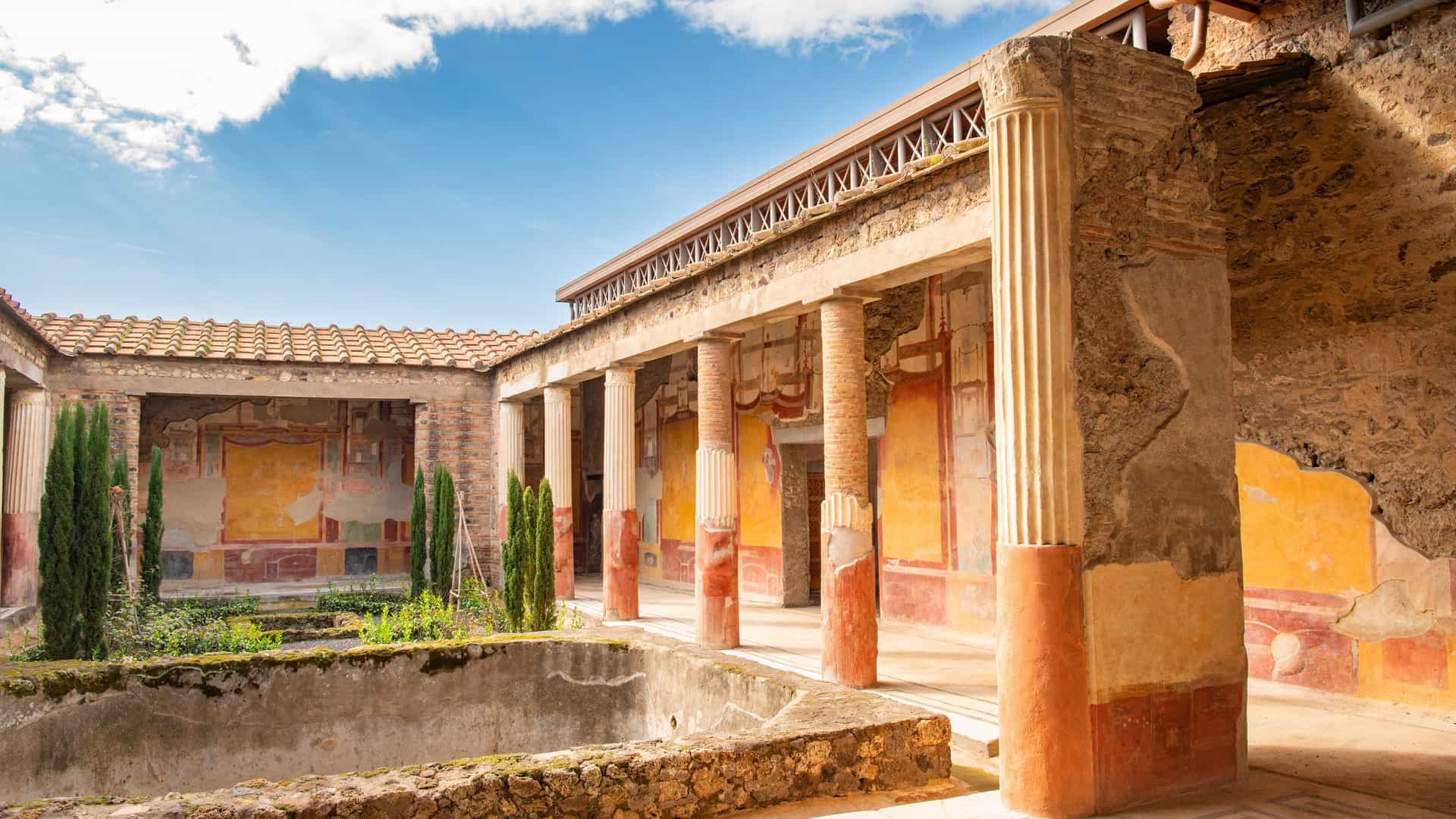 A courtyard of an ancient Roman villa in the ruins of Pompeii, Italy, showing a rectangular pool, a covered walkway with a line of columns, and frescoes on the walls.