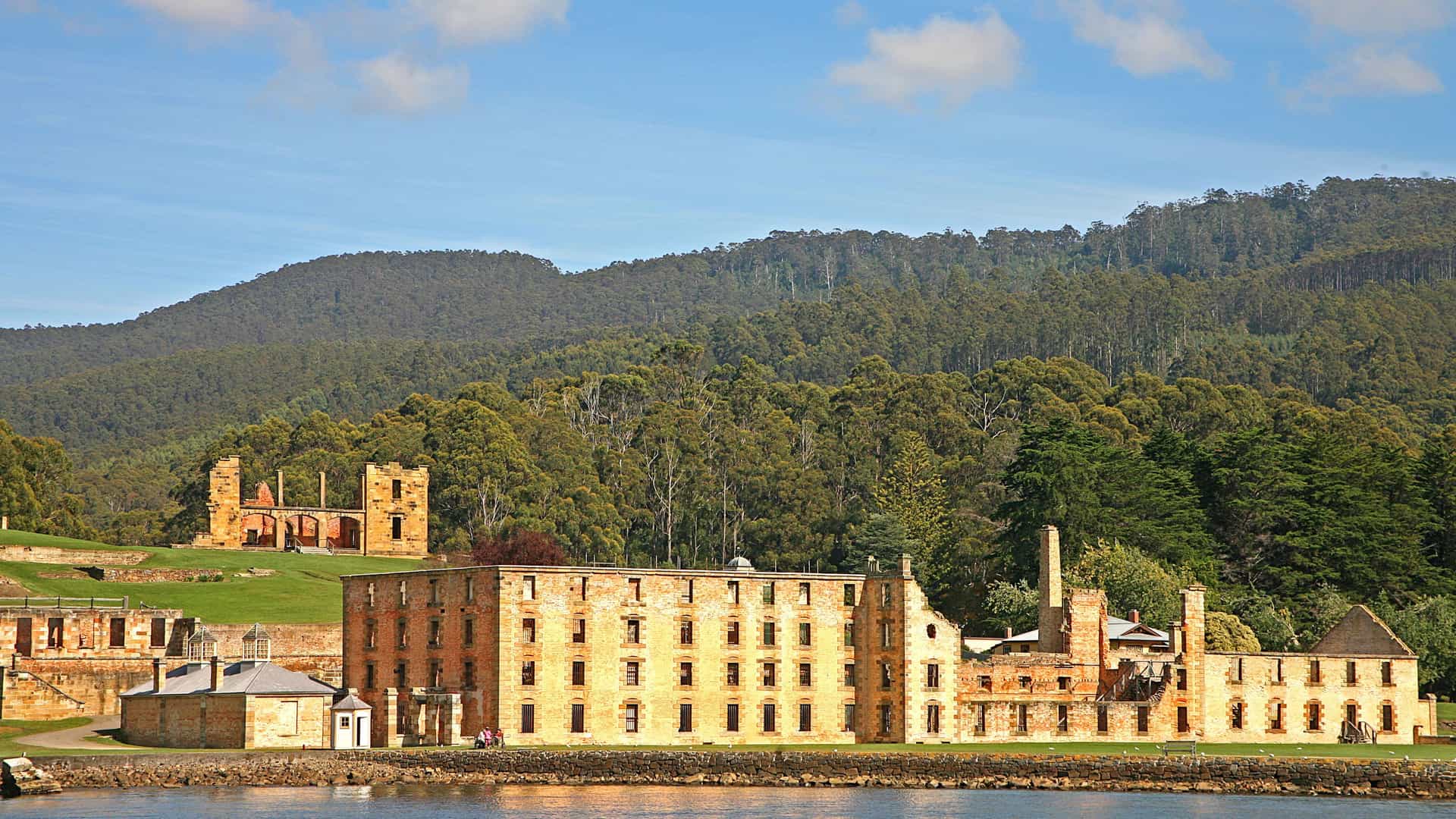 A panoramic view of the historic Port Arthur penal colony ruins in Tasmania, Australia, with multiple stone buildings and structures facing the water and a lush, tree-covered hillside in the background.