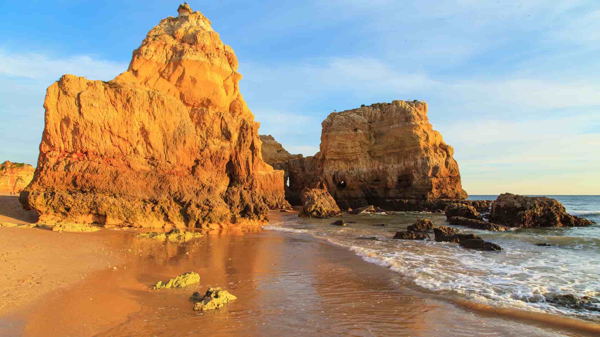 A golden-hour shot of a sandy beach in Portimão, Portugal, with the clear water of the Atlantic Ocean gently washing over the sand and the large, jagged limestone rock formations of the Algarve coast lit by the sun.