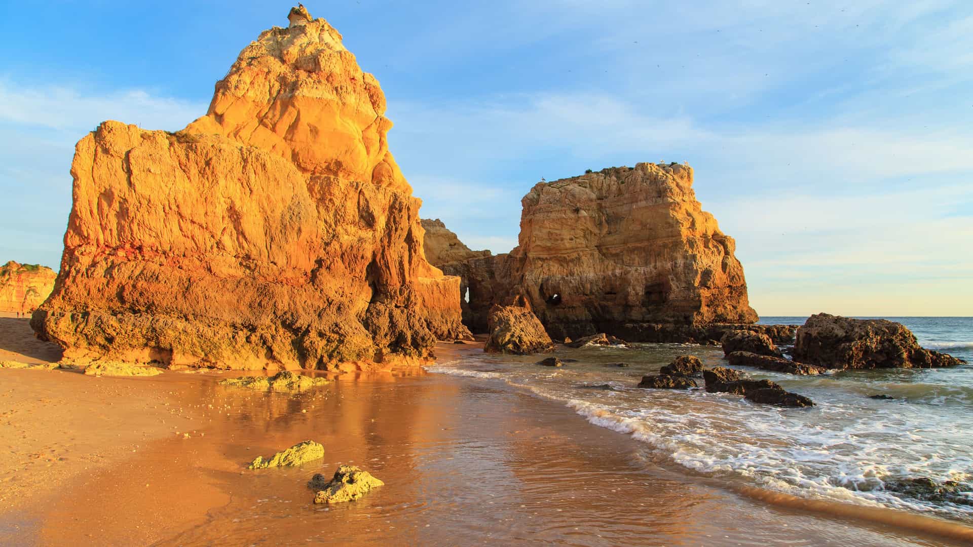 A golden-hour shot of a sandy beach in Portimão, Portugal, with the clear water of the Atlantic Ocean gently washing over the sand and the large, jagged limestone rock formations of the Algarve coast lit by the sun.