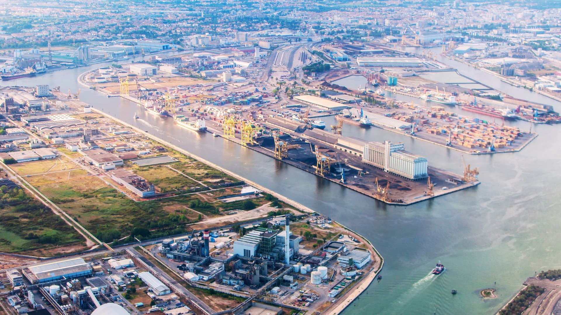 An aerial view of the Porto Marghera industrial complex and oil refinery near Venice, Italy, showing a vast network of pipelines, storage tanks, towering structures, and industrial buildings against the backdrop of the port and water channels.