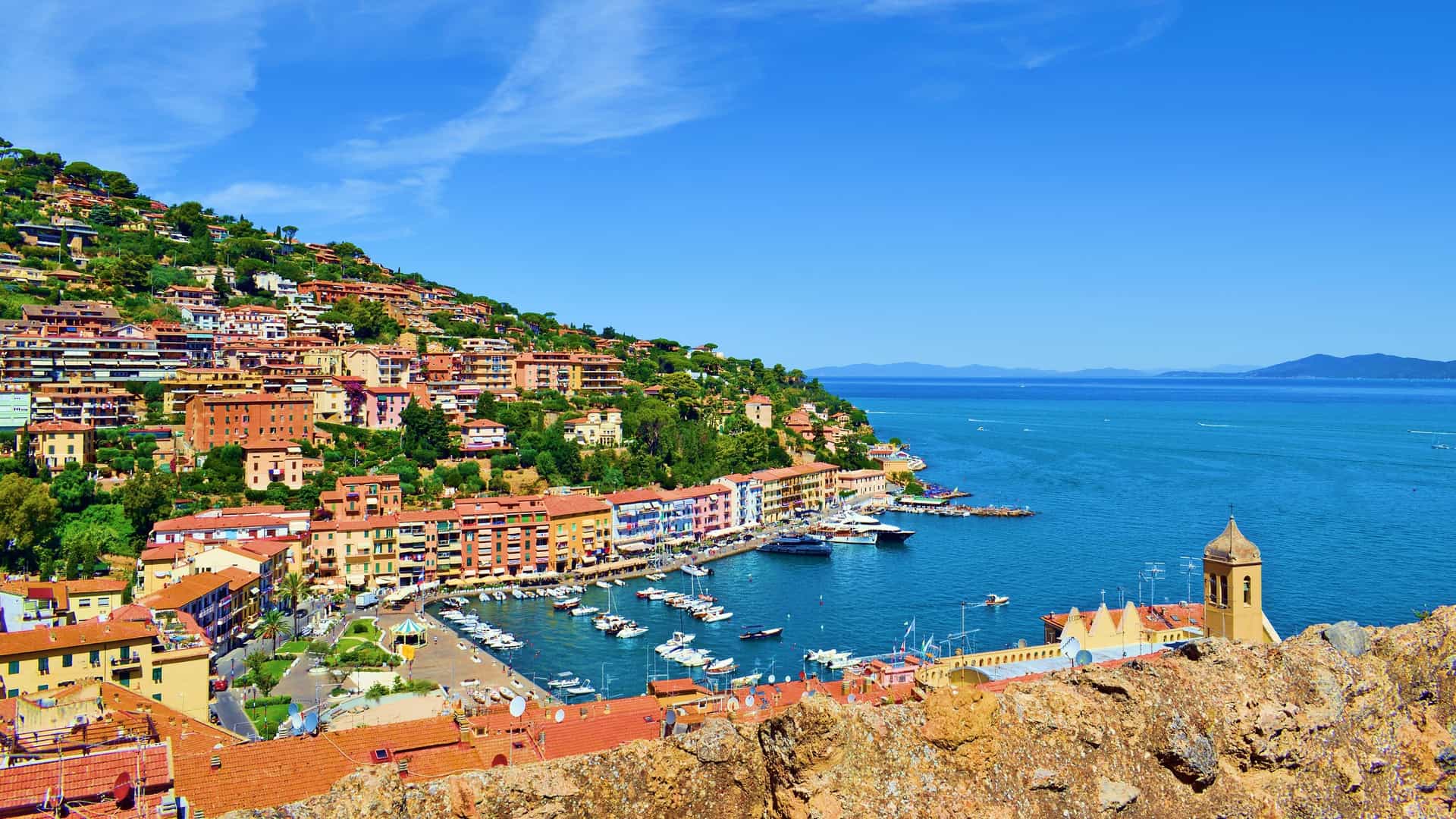 A panoramic view of Porto Santo Stefano, Italy, with its colorful houses built on a steep hillside overlooking the marina and the sparkling Tyrrhenian Sea.