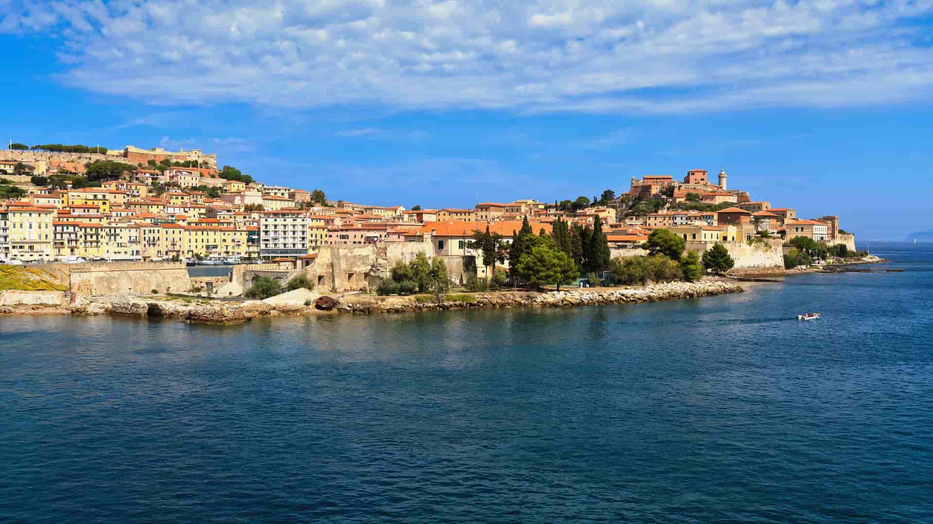 A beautiful wide shot of the Port of Portoferraio on Elba Island, Italy, showcasing the historic town's terracotta-roofed buildings and fortifications sprawling up the hillside from the blue sea.