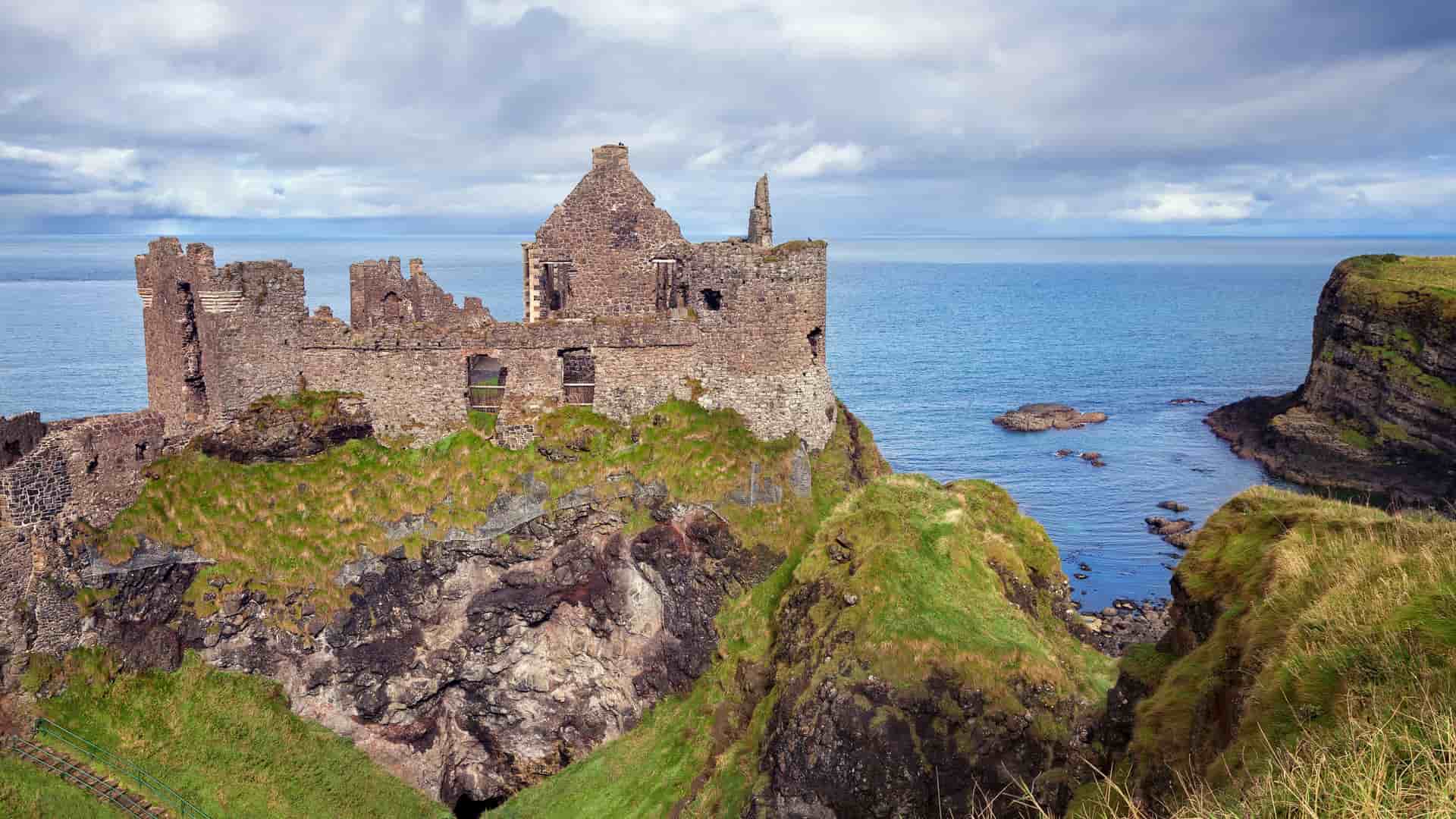A stunning photograph of the medieval Dunluce Castle ruins, perched on a dramatic cliff overlooking the North Atlantic Ocean near Portrush in Northern Ireland.