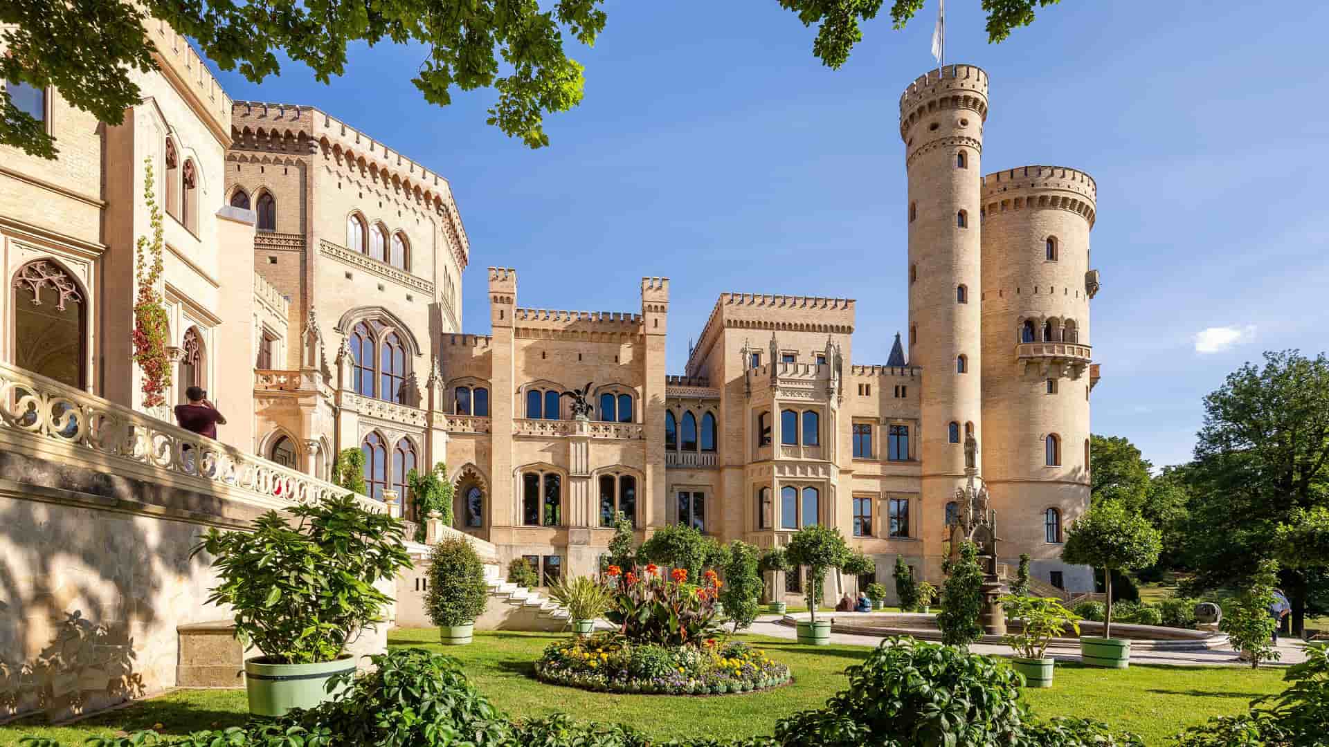 The ornate facade of the Babelsberg Palace in Potsdam, Germany, featuring its grand towers and elegant windows. The light-colored castle is surrounded by a beautifully manicured garden with a circular flower bed and well-trimmed trees, all under a clear blue sky.