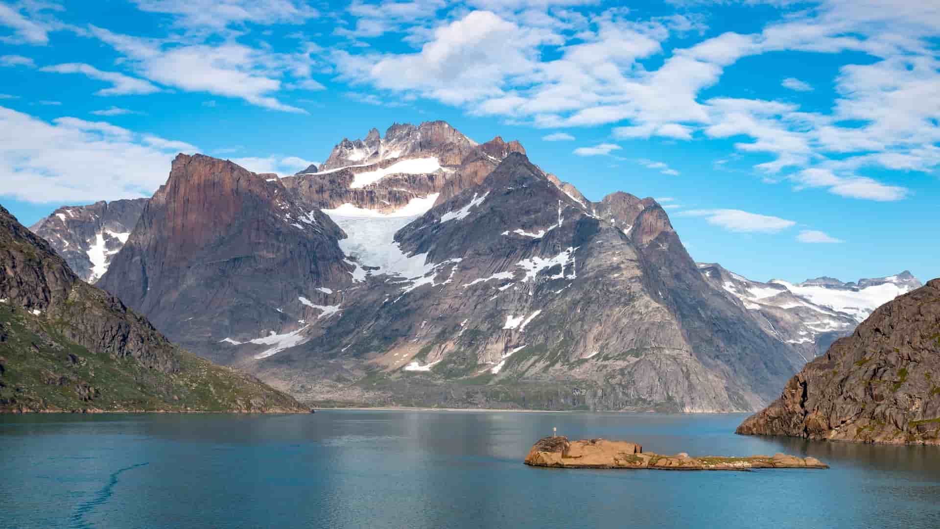 A beautiful panoramic shot of the Prince Christian Sound, Greenland, with massive, snow-capped mountains and glaciers towering over a small rocky island in the tranquil fjord.