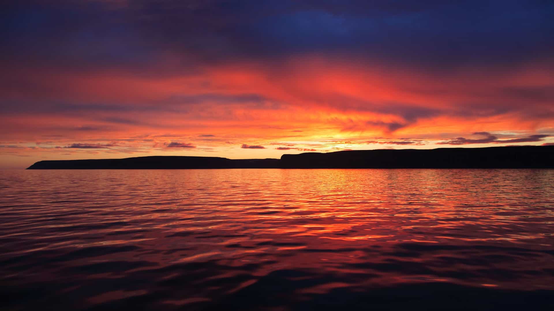 A beautiful, dramatic sunset over Prince Leopold Island, Nunavut, Canada, with a vibrant sky casting a fiery red and orange reflection on the calm water.