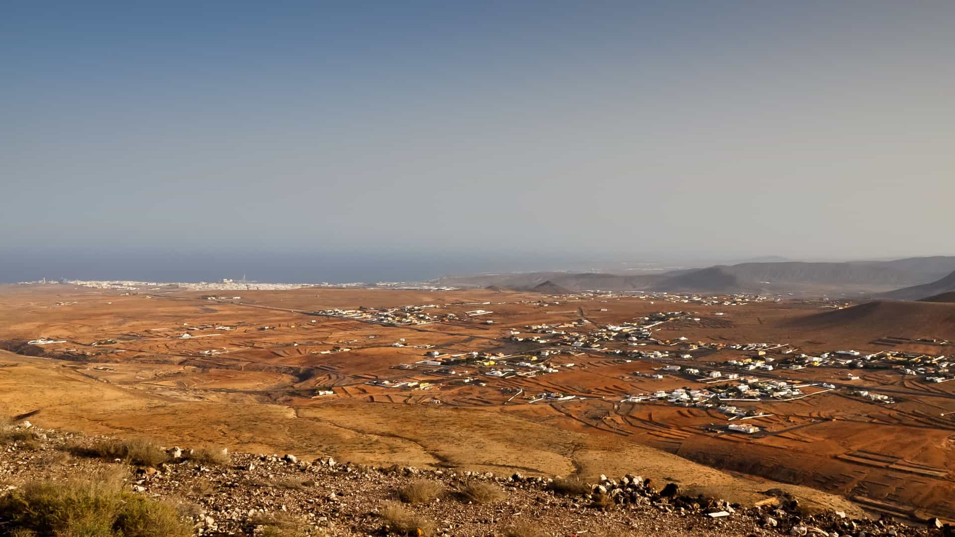 A panoramic aerial shot of the arid landscape of Fuerteventura, Spain, with the town of Antigua visible in the foreground and the Atlantic Ocean in the background.