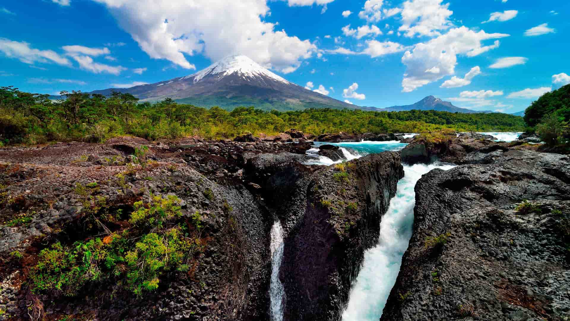 A wide shot of the spectacular PetrohuƩ Falls near Puerto Montt, Chile, with turquoise water cascading over volcanic rock, set against the backdrop of the snow-capped Osorno Volcano.