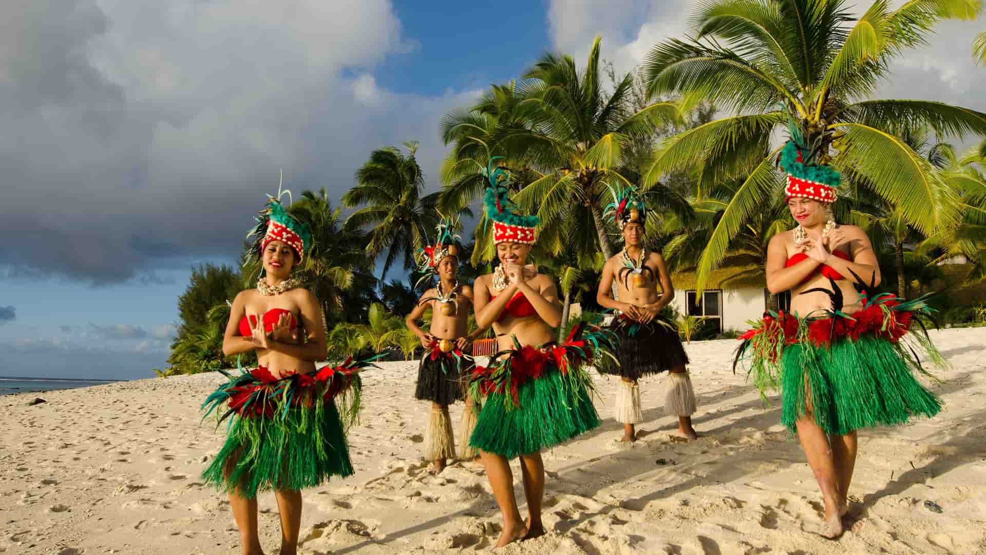 A group of five Cook Islands women in traditional dress with green skirts, red feathers, and colorful headdresses perform a dance on a sandy Rarotonga beach surrounded by palm trees.