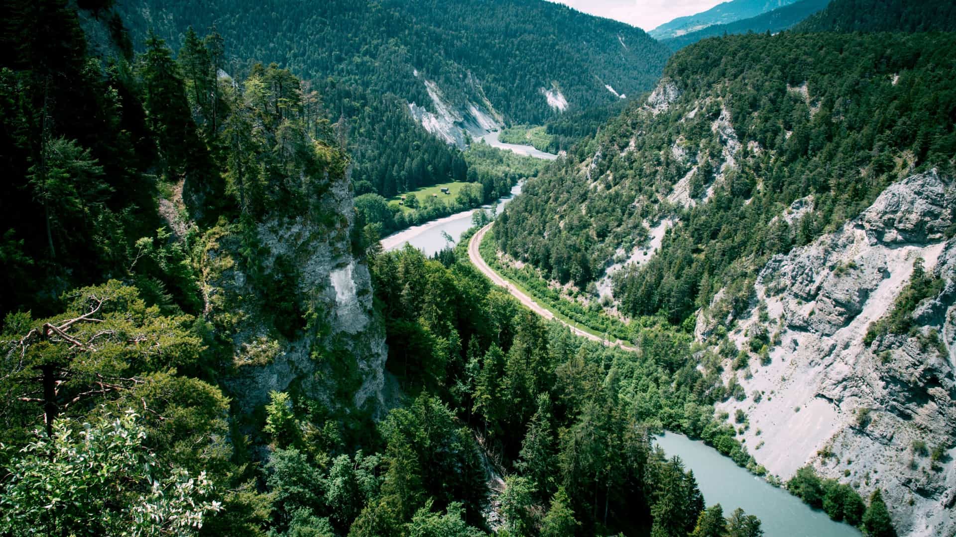 An aerial view of the Rhine Gorge, Switzerland, with a winding river and a small road carving through a lush, green forest and steep, rocky cliffs.