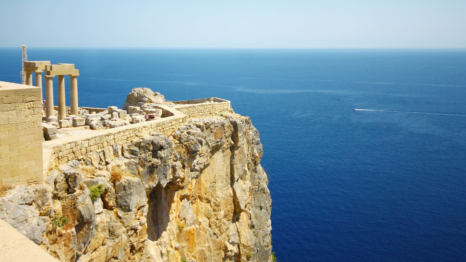 A scenic view of the Acropolis of Lindos ruins perched on a cliffside overlooking the deep blue Mediterranean Sea and a clear sky on the island of Rhodes, Greece.