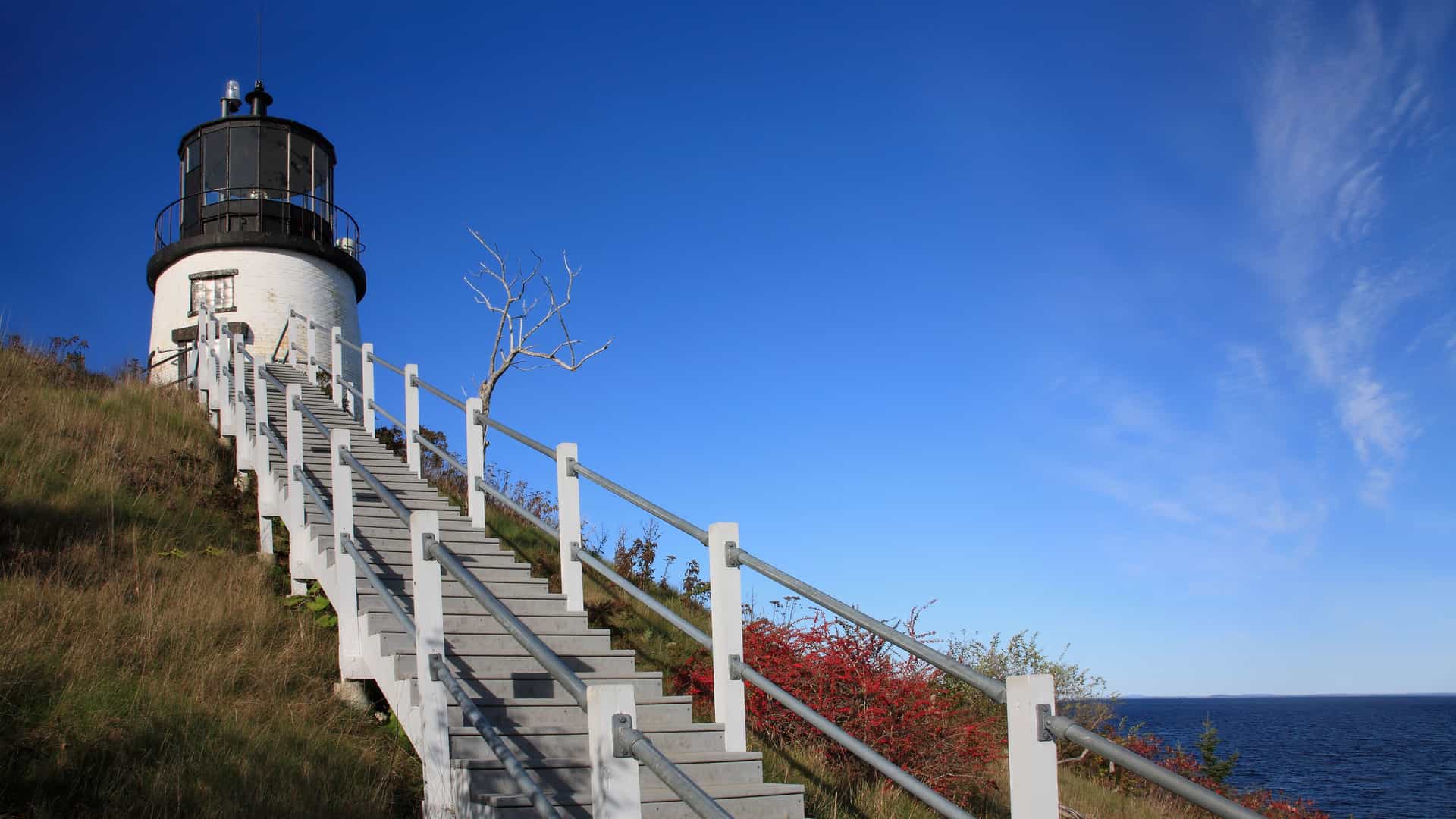 A view of the Owl's Head Lighthouse in Rockland, Maine, with a long staircase leading up to the historic white lighthouse, set against a bright blue sky and the Atlantic Ocean.