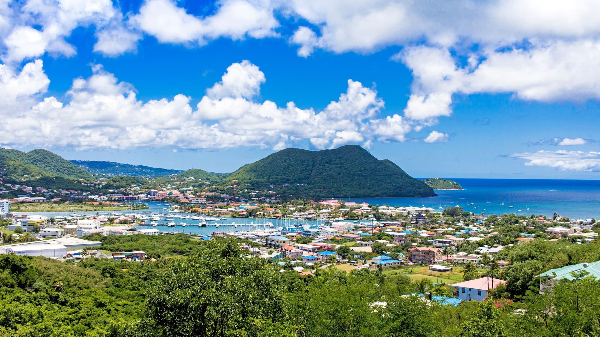 A stunning panoramic view of Rodney Bay on the island of St. Lucia, showing a bustling harbor filled with sailboats, a colorful town, and lush green hills under a bright blue, cloudy sky.