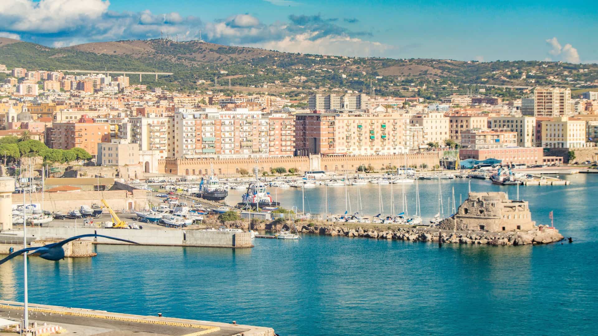 A beautiful view of the harbor and waterfront of Civitavecchia, Italy, with docked boats, historic buildings, and lush hills in the background.