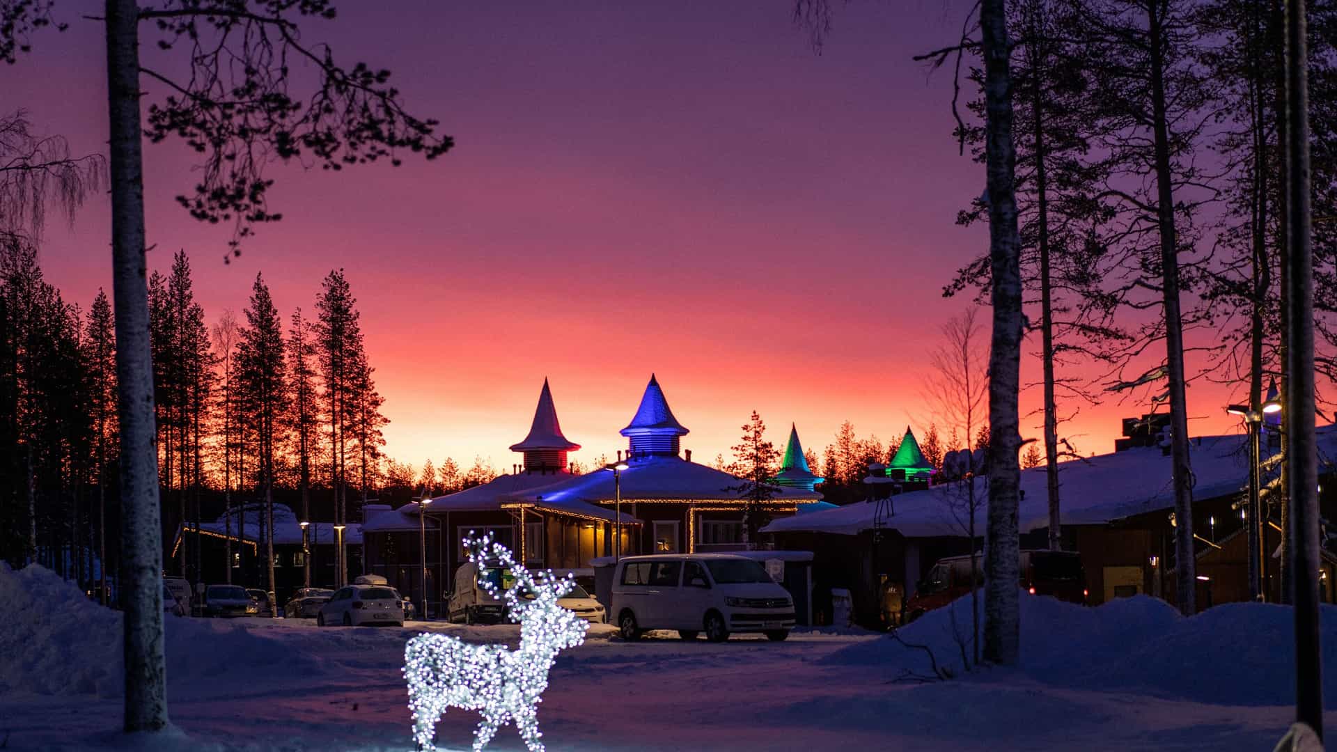 "A beautiful winter scene at the Santa Claus Village in Rovaniemi, Finland, with festive lights and a reindeer statue, set against a colorful sunset sky. "