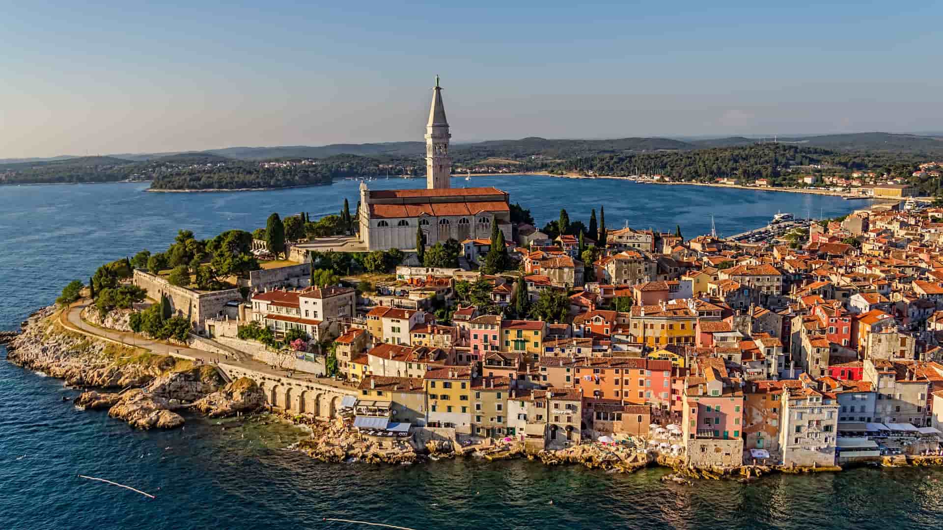 An aerial view of the historic town of Rovinj, Croatia, with its colorful buildings and the bell tower of St. Euphemia's Church dominating the skyline, all perched on a peninsula jutting into the Adriatic Sea.