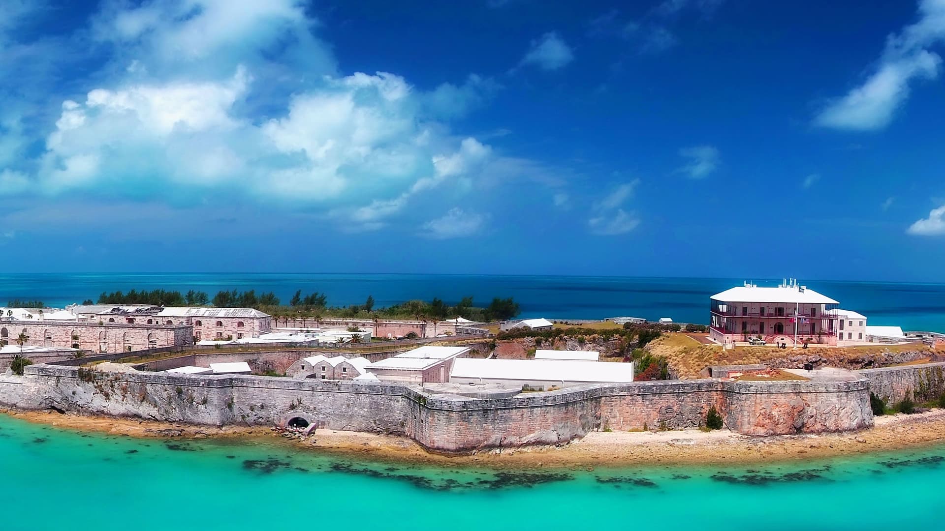 An aerial view of the Royal Naval Dockyard in Bermuda, with a cruise ship docked at the pier, surrounded by a historic stone fortress, buildings, and the turquoise waters of the Atlantic Ocean.