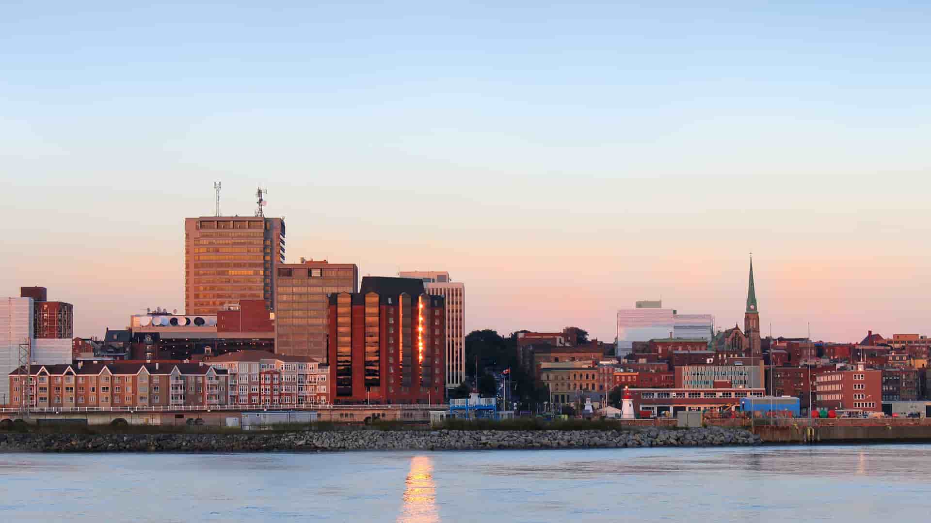 A cityscape view of a small town on the Saguenay River with a mix of modern high-rises and historic buildings with a church steeple, all bathed in the warm light of dusk.