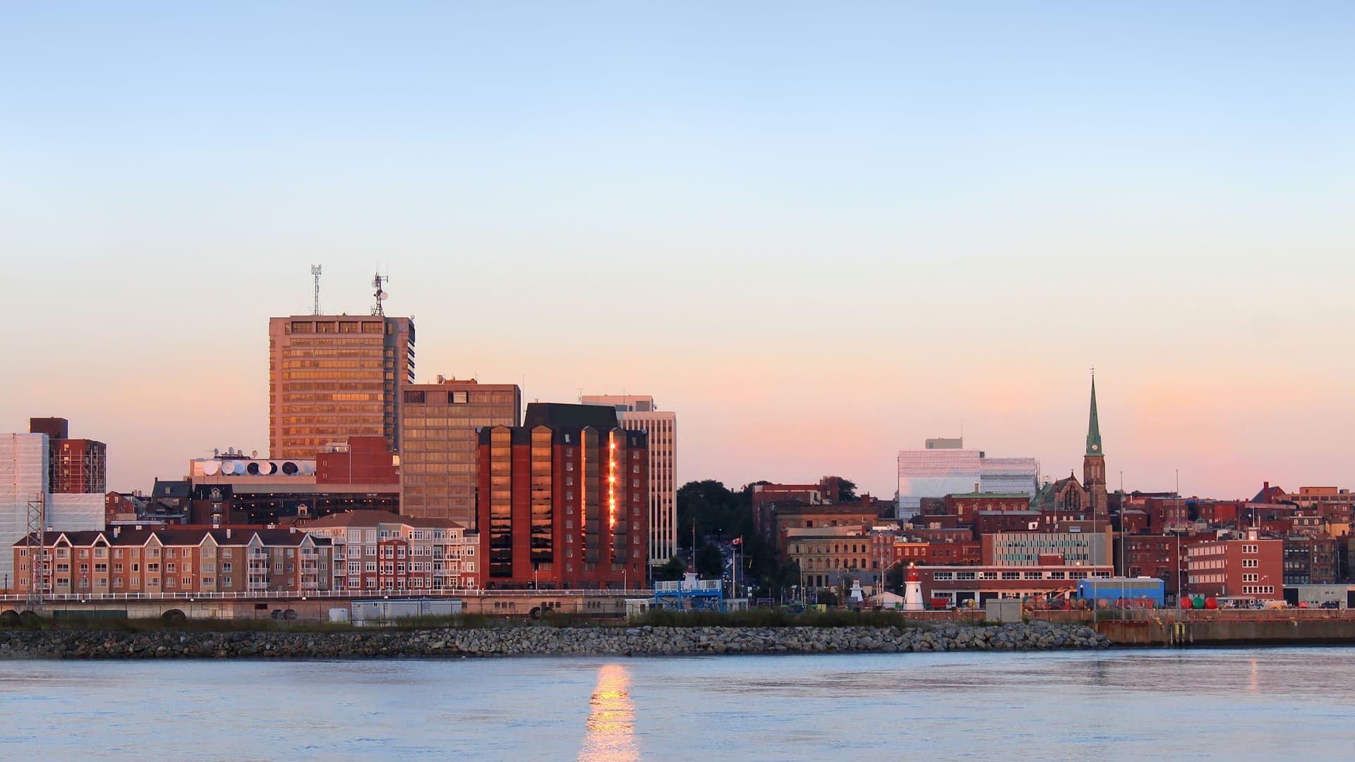 A cityscape view of a small town on the Saguenay River with a mix of modern high-rises and historic buildings with a church steeple, all bathed in the warm light of dusk.