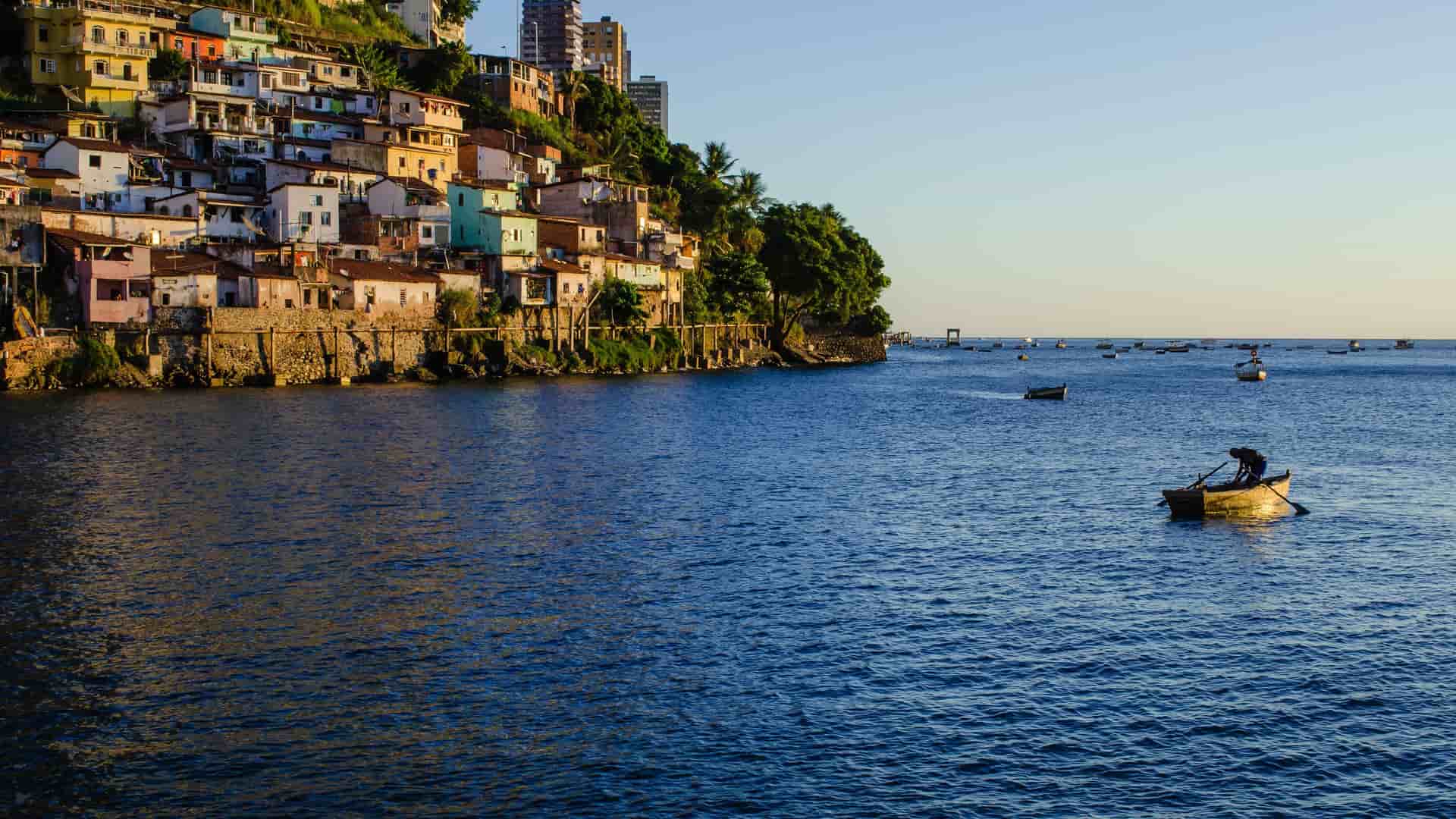 A vibrant waterside view of Salvador de Bahia, Brazil, featuring a hillside covered in colorful houses overlooking the calm blue waters of All Saints' Bay, where a person is rowing a small boat.