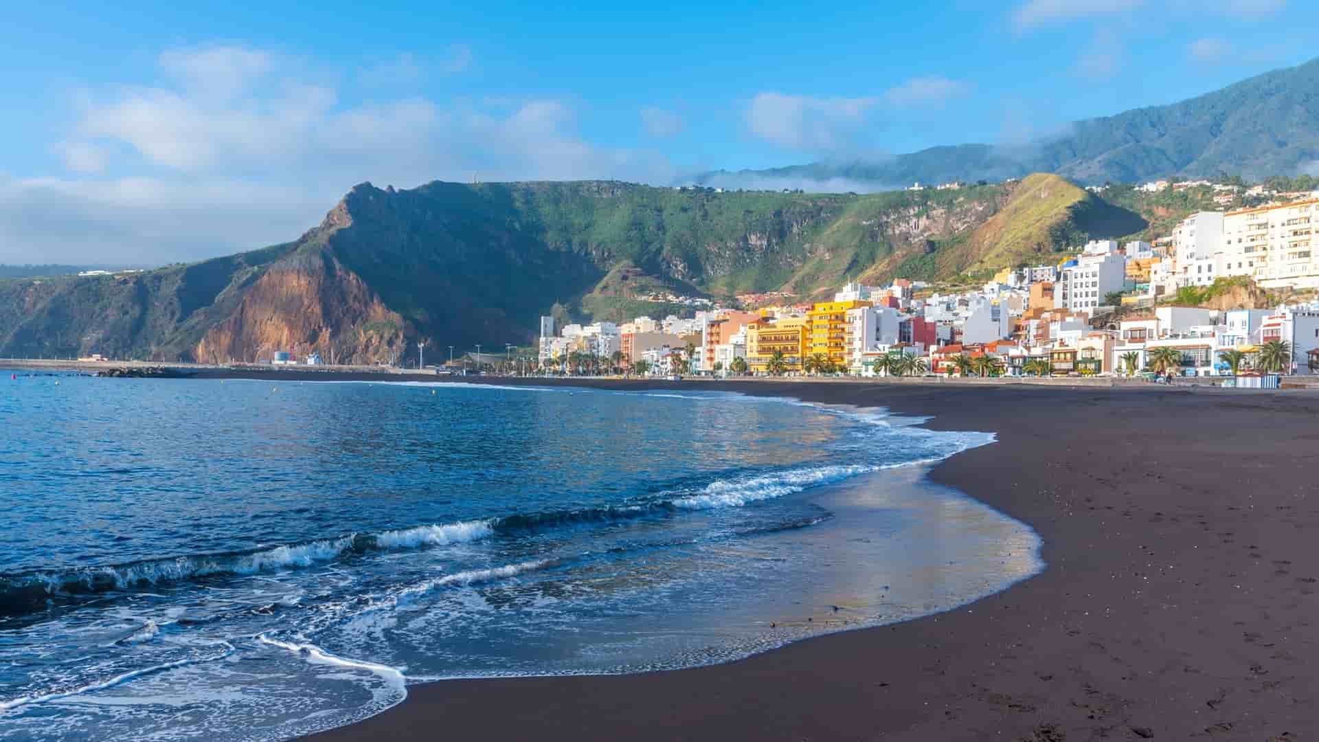 A stunning view of Santa Cruz de la Palma's black sand beach, with gentle waves from the Atlantic Ocean, a colorful town, and green, volcanic mountains in the background.