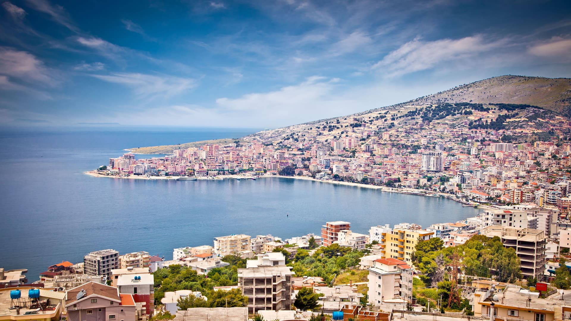 A scenic daytime shot of Sarandë, Albania, a coastal city with white buildings clinging to a hillside overlooking the clear blue Ionian Sea.